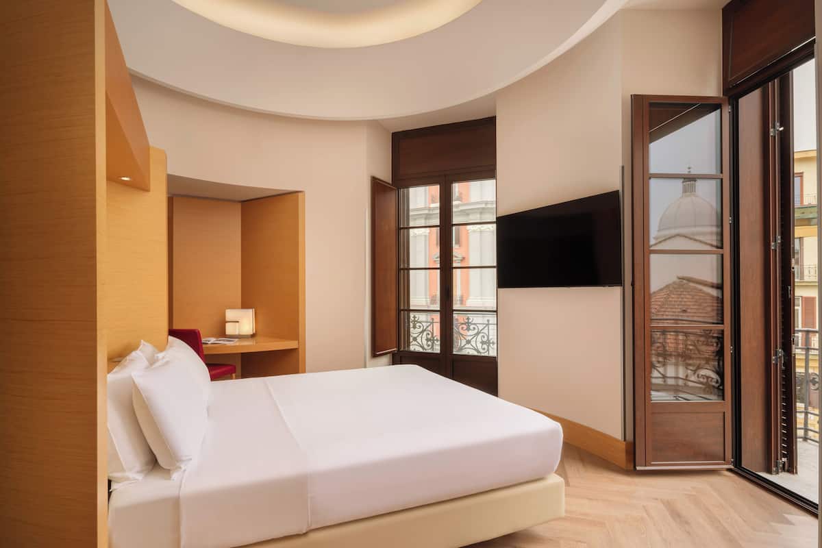 Een hotelkamer met een bed, een bureau, een stoel, een televisie en een balkon met uitzicht op gebouwen.