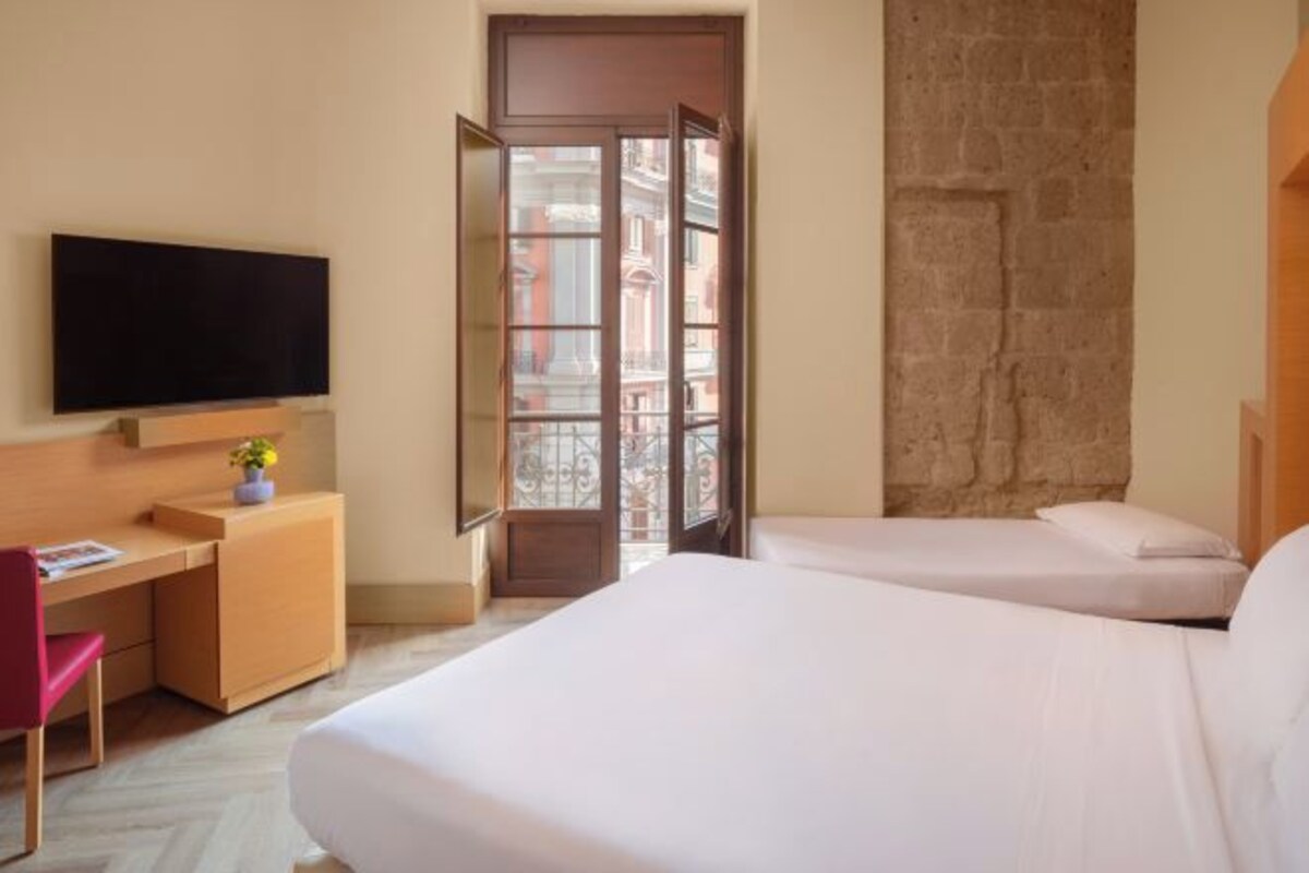 A hotel room with a bed, a desk, a chair, a TV, and a stone wall.