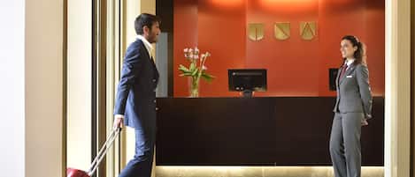 A reception area with a reception desk, a vase with flowers, and a man pulling a suitcase.