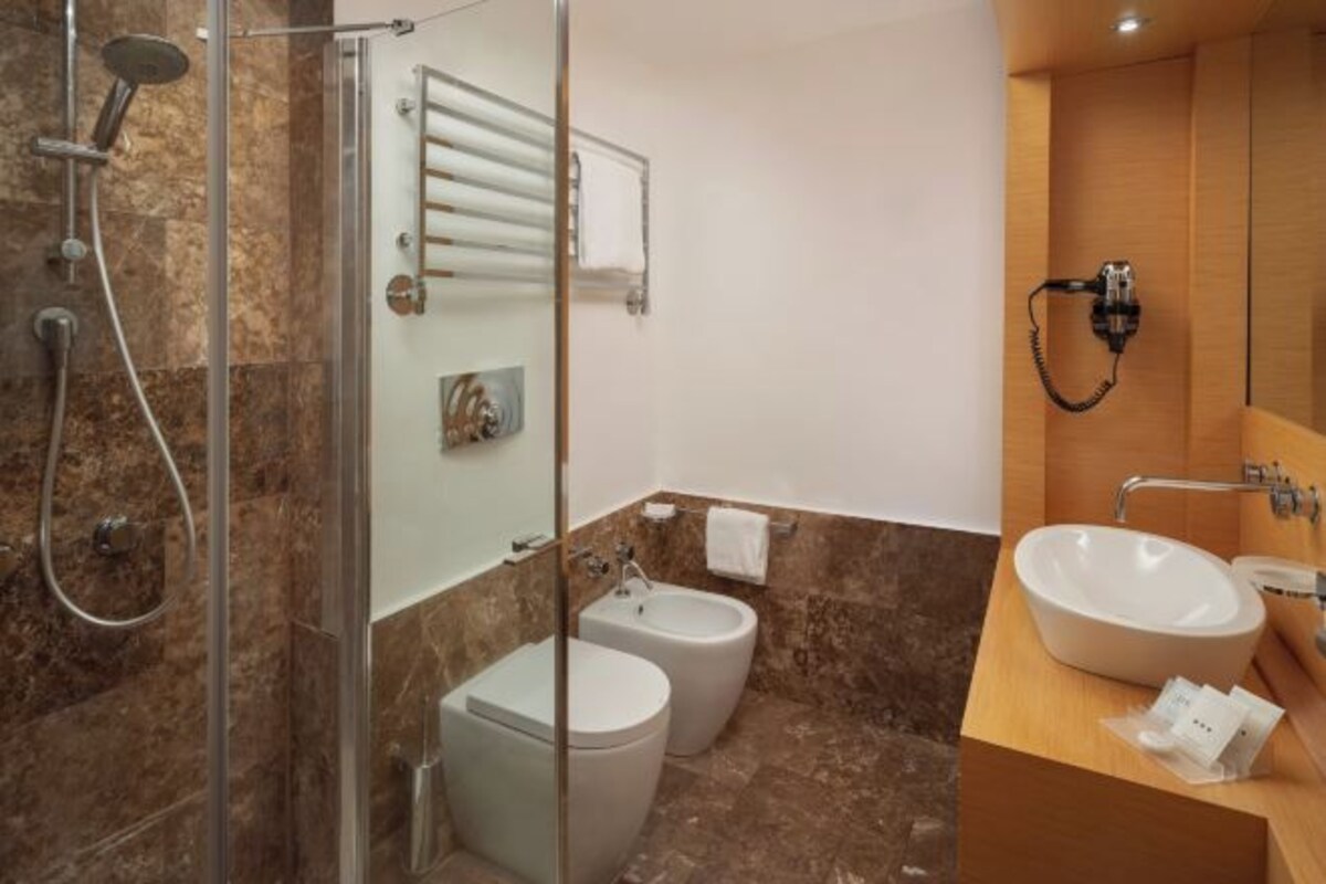 A bathroom with a glass-enclosed shower, a bidet, a toilet, and a sink with a towel dispenser.