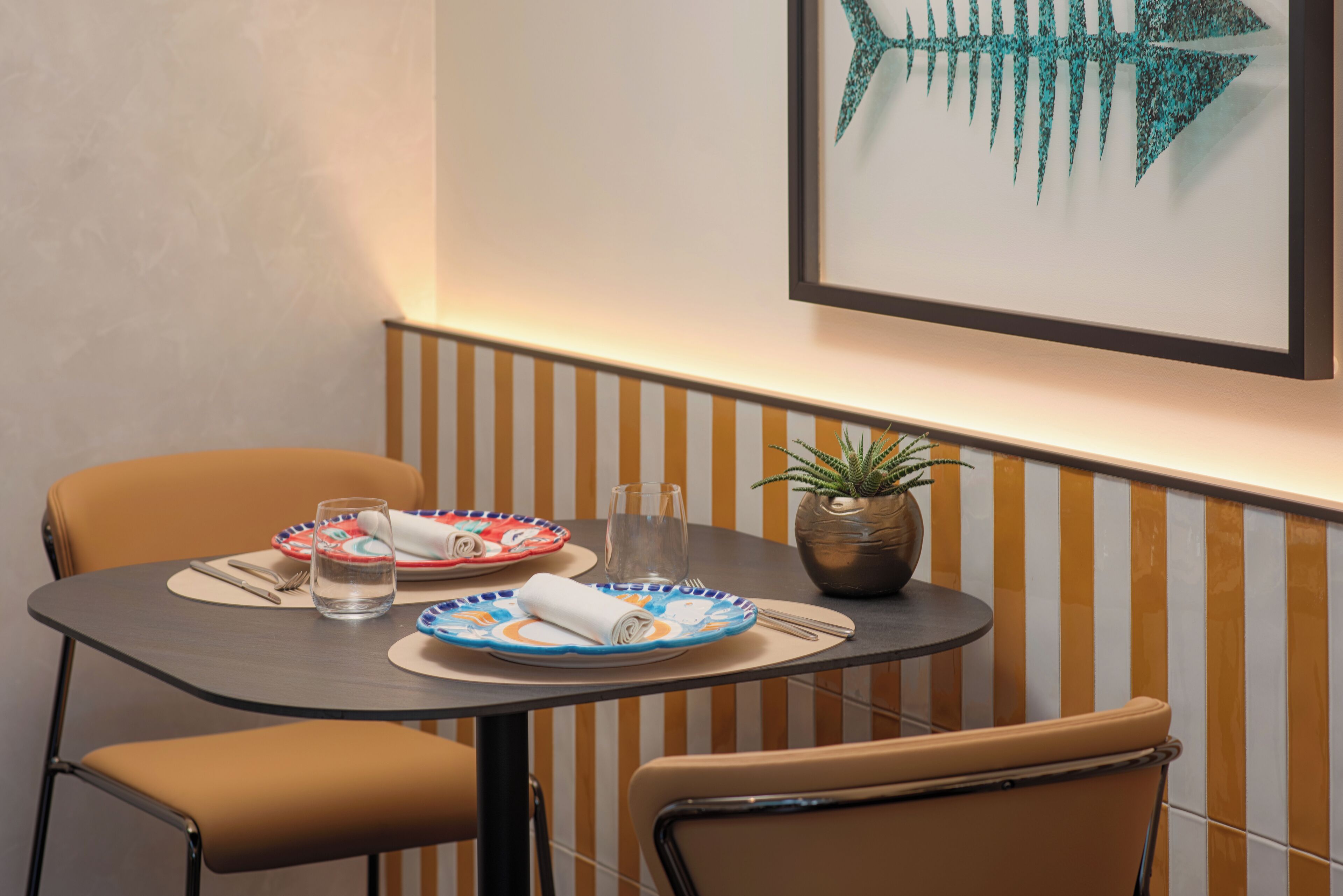 A dining area with a table set for two, a potted plant, and a framed artwork on the wall.