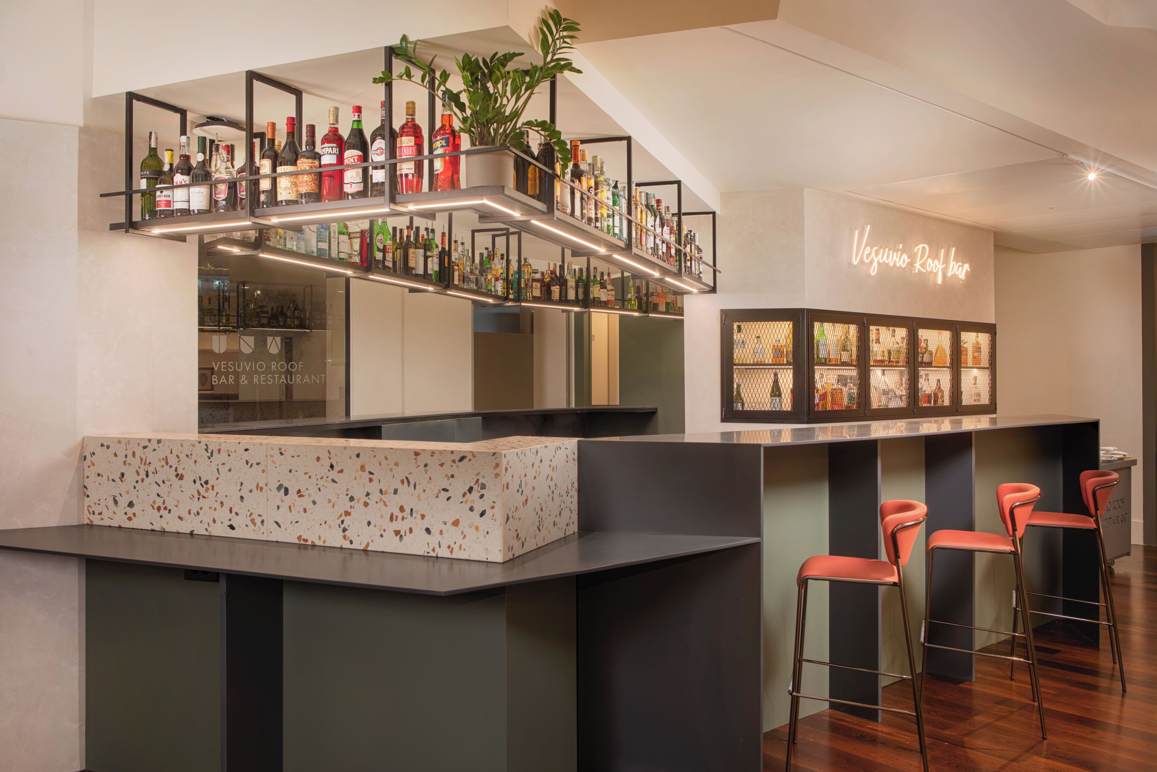 A bar area with a counter, stools, shelves with bottles, and a neon sign.