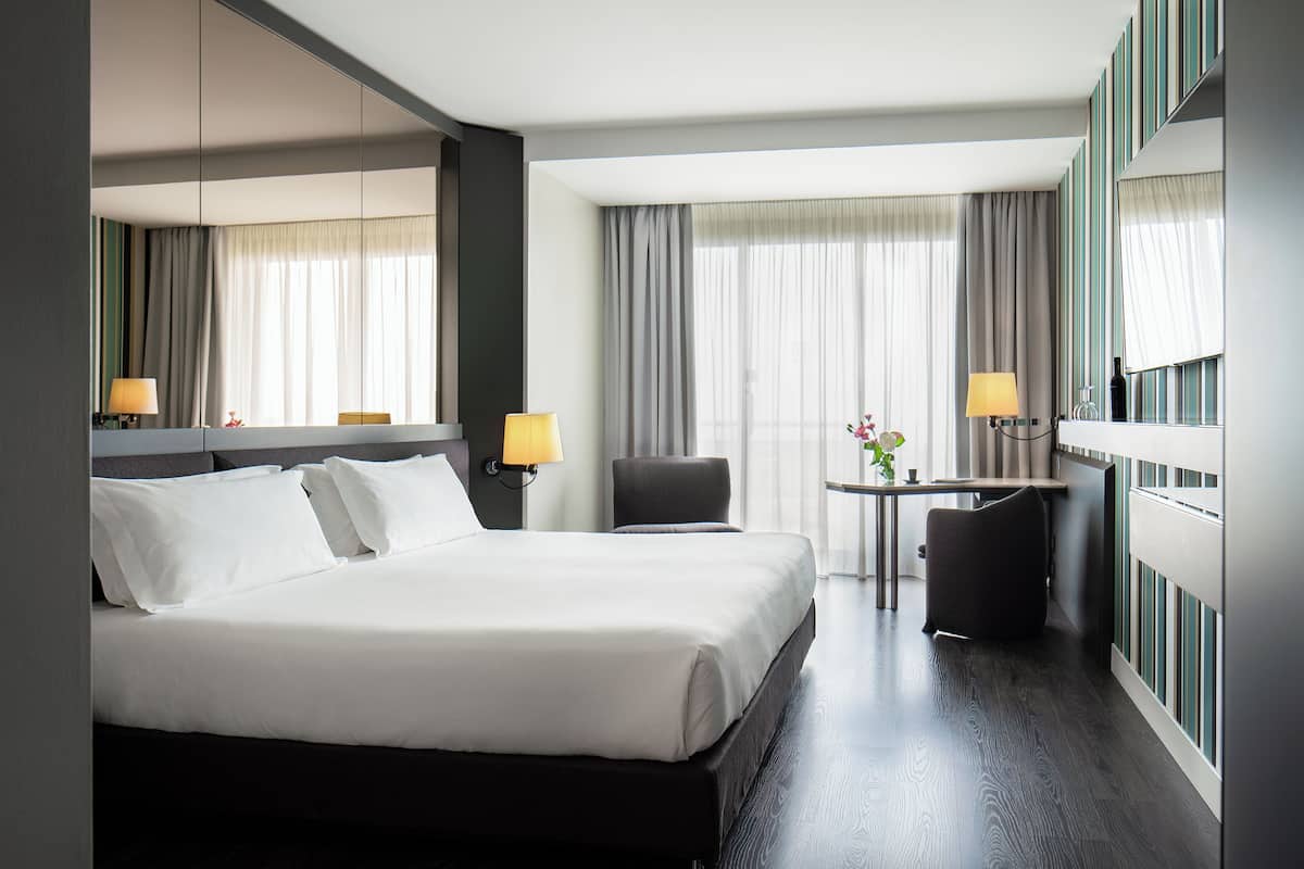 Una habitación de hotel moderna con una cama grande, un escritorio, una silla y vistas a la ciudad desde las ventanas.
