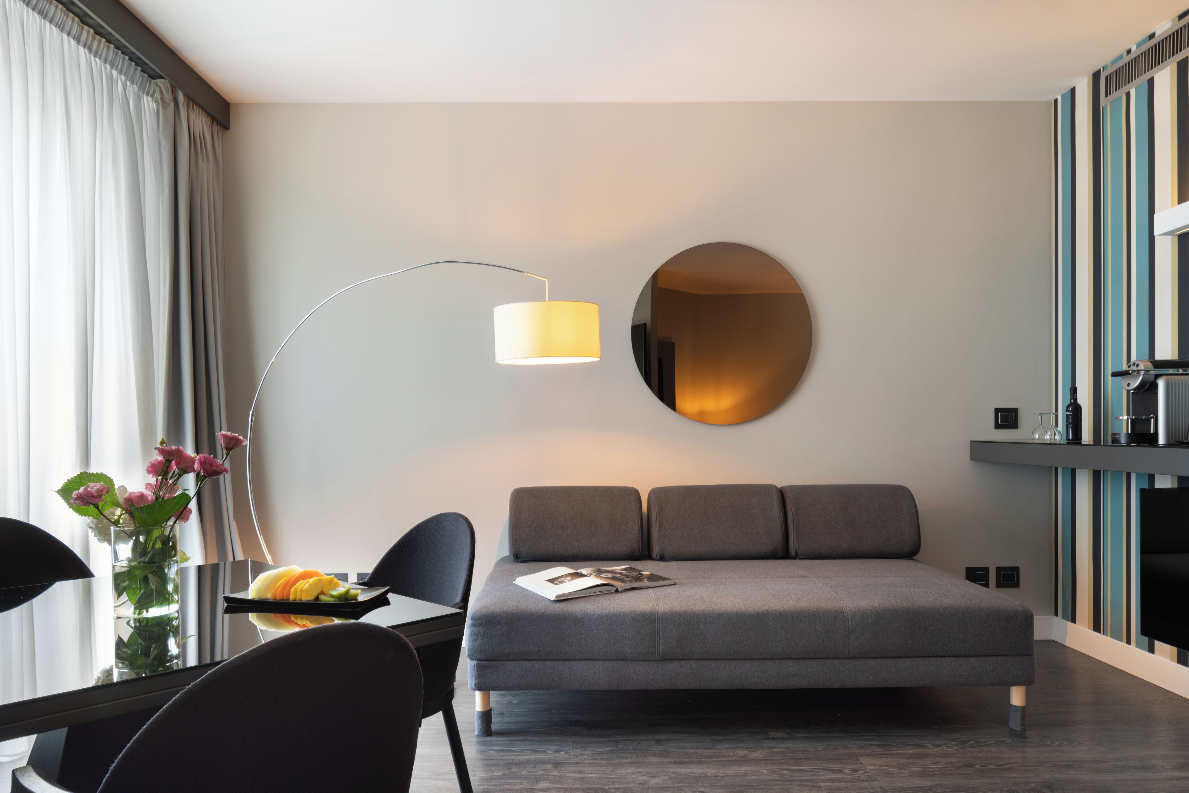 Una habitación de hotel moderna con un sofá gris, un espejo redondo, una lámpara de pie, una mesa de comedor con sillas y un jarrón con flores.