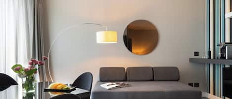 A modern hotel room with a grey sofa, a round mirror, a floor lamp, a dining table with chairs, and a vase with flowers.