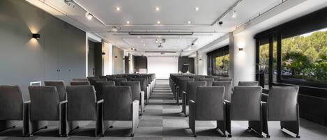 A conference room with rows of chairs, ceiling lights, and a projector screen.