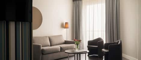 A modern hotel room with a sofa, armchair, and a small table with a vase of flowers.