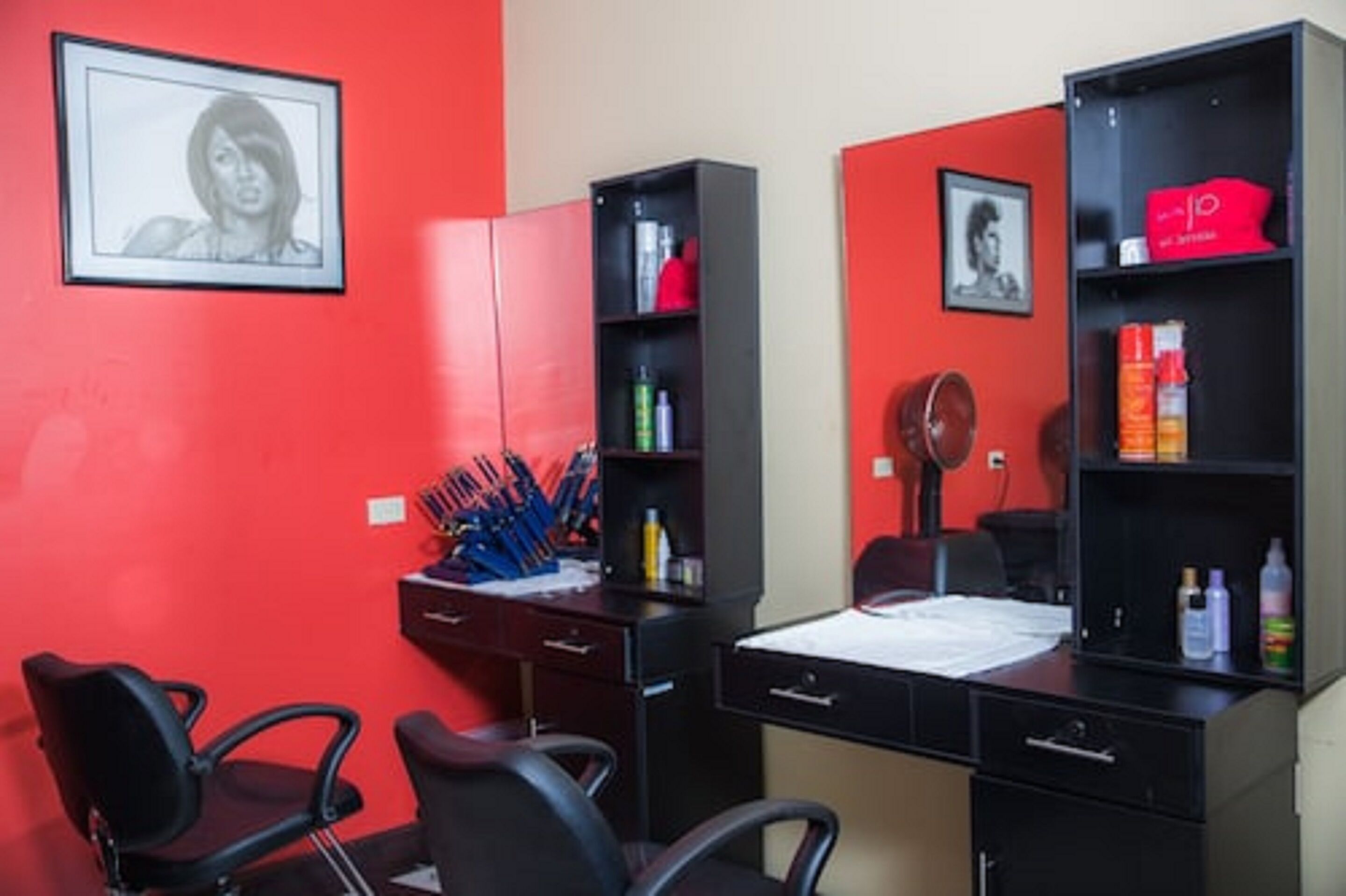 A room with a red wall, a black desk, two black chairs, a bed, and a bookshelf with various items.