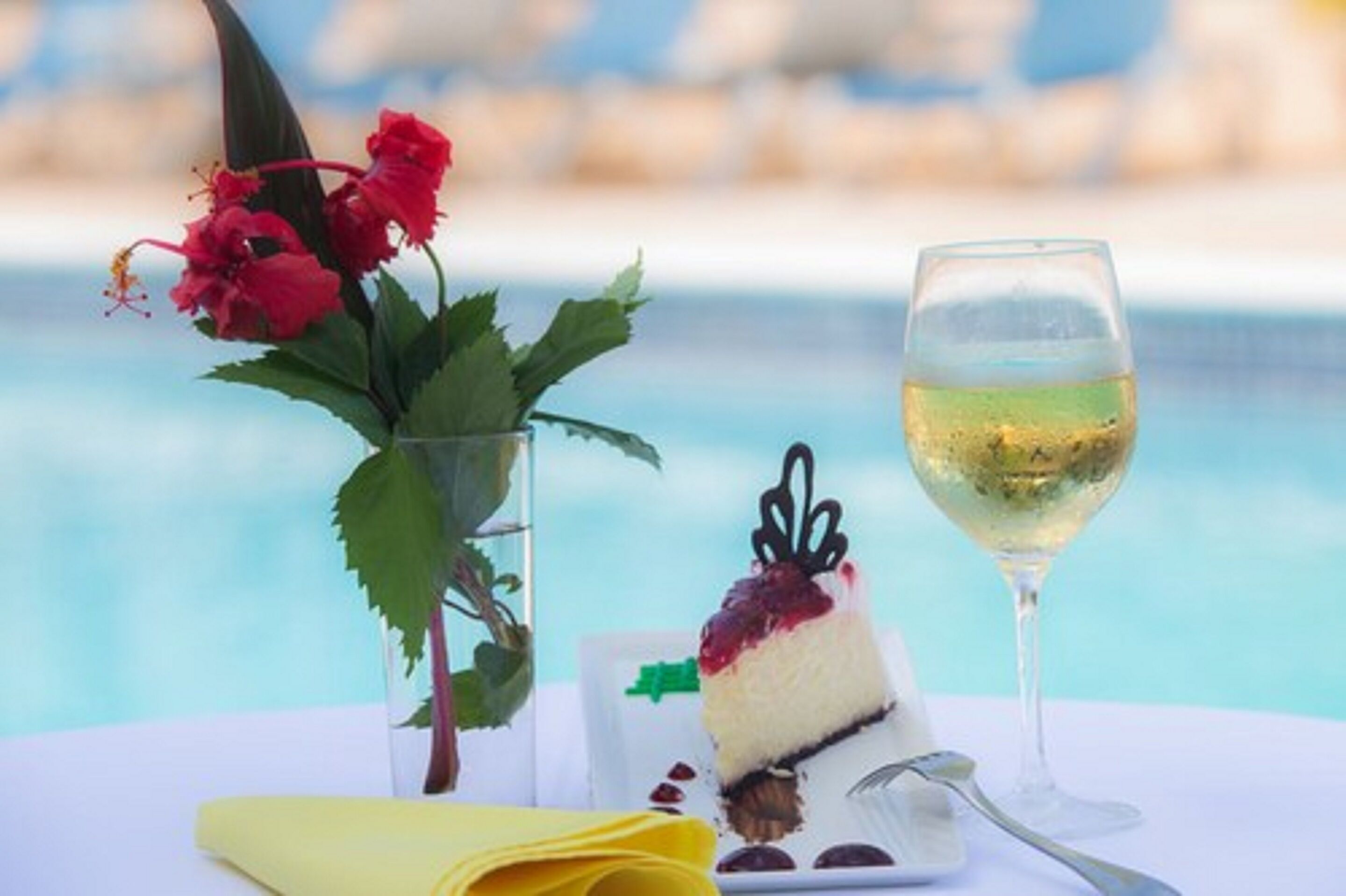 Un postre con una porción de pastel y una copa de vino blanco sobre una mesa junto a una piscina.
