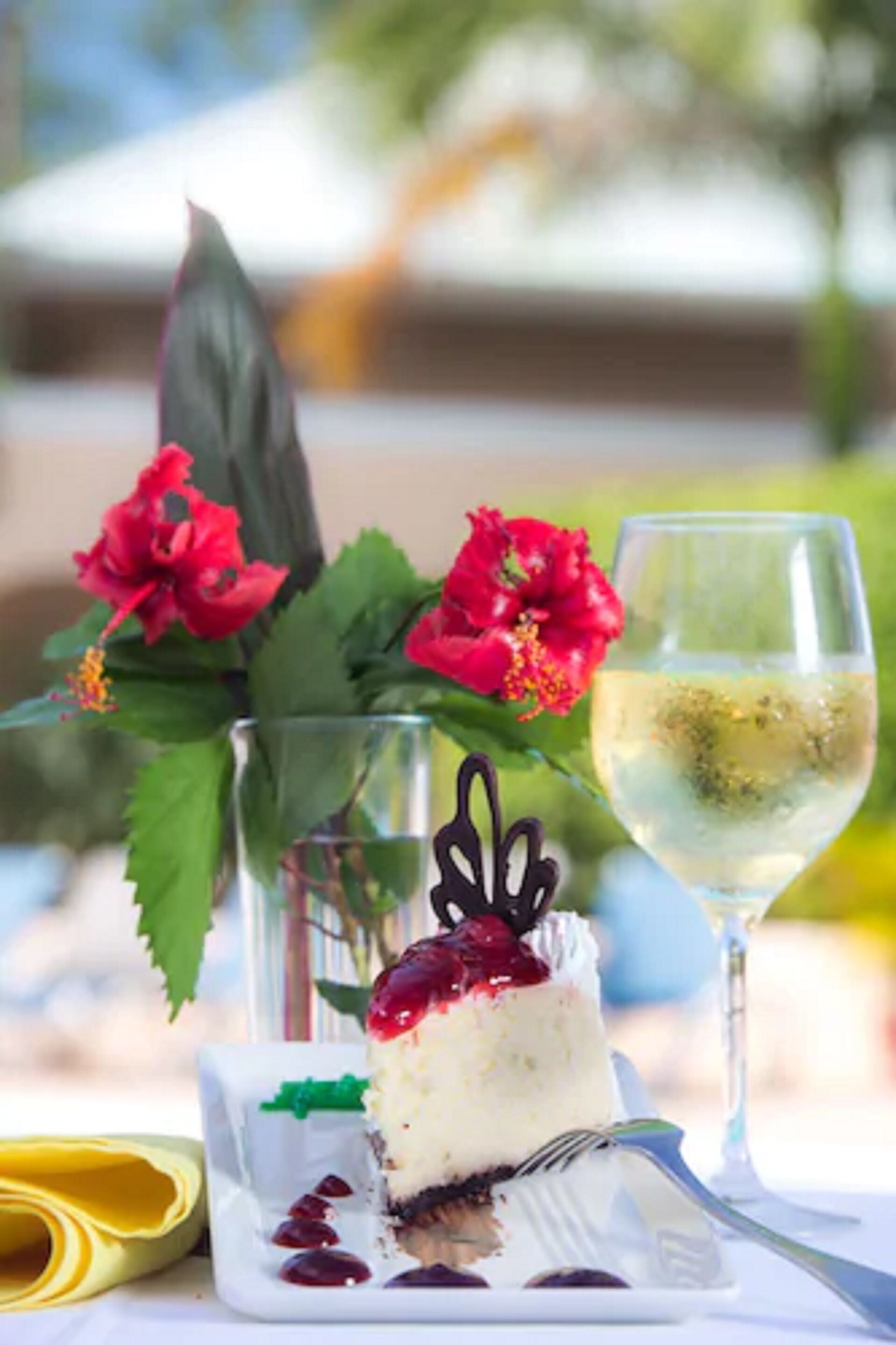 A dessert with a slice cut out, a glass of white wine, and red flowers in the background.