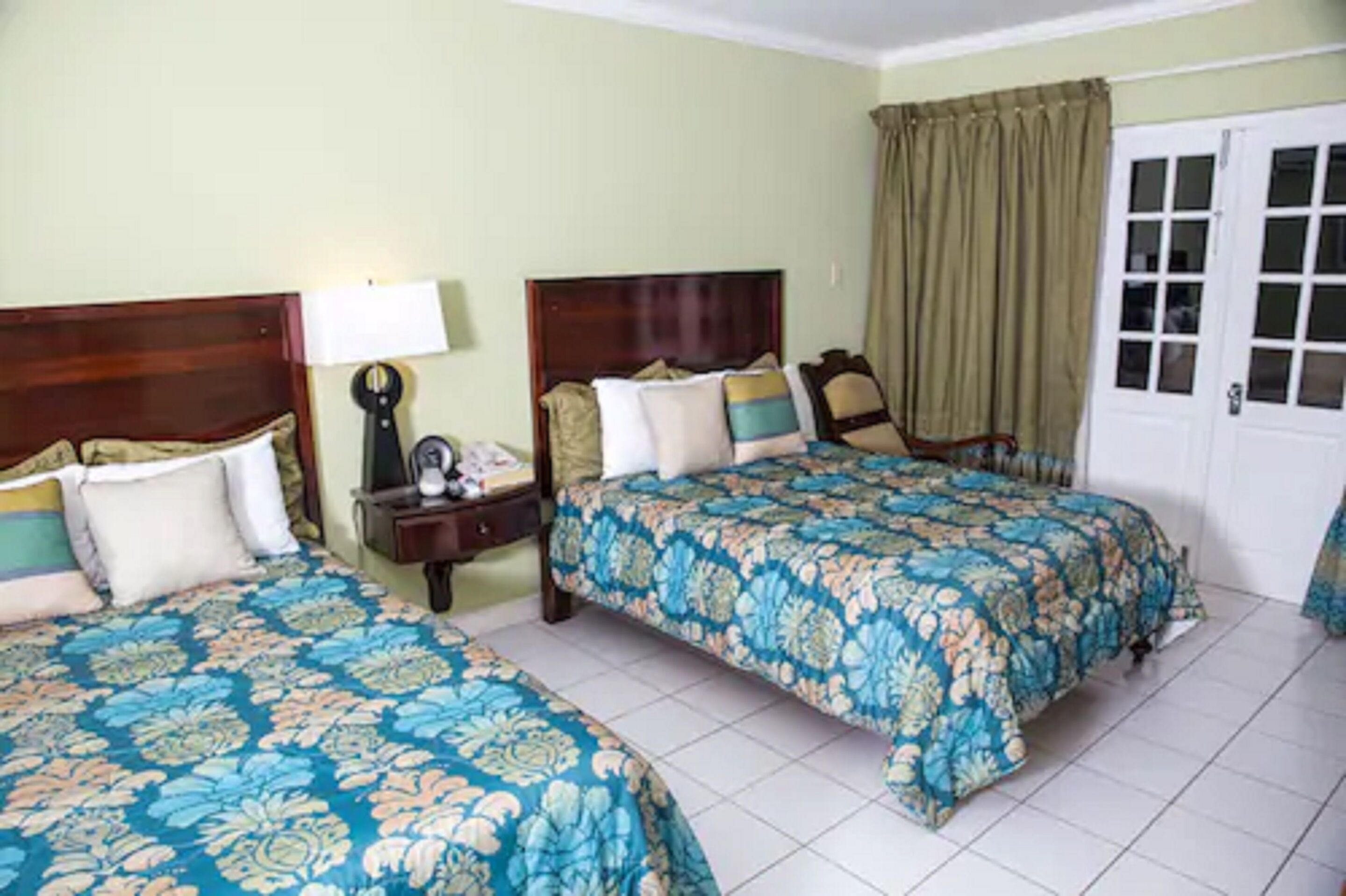 A hotel room with two beds, a nightstand, a lamp, and a door with curtains.
