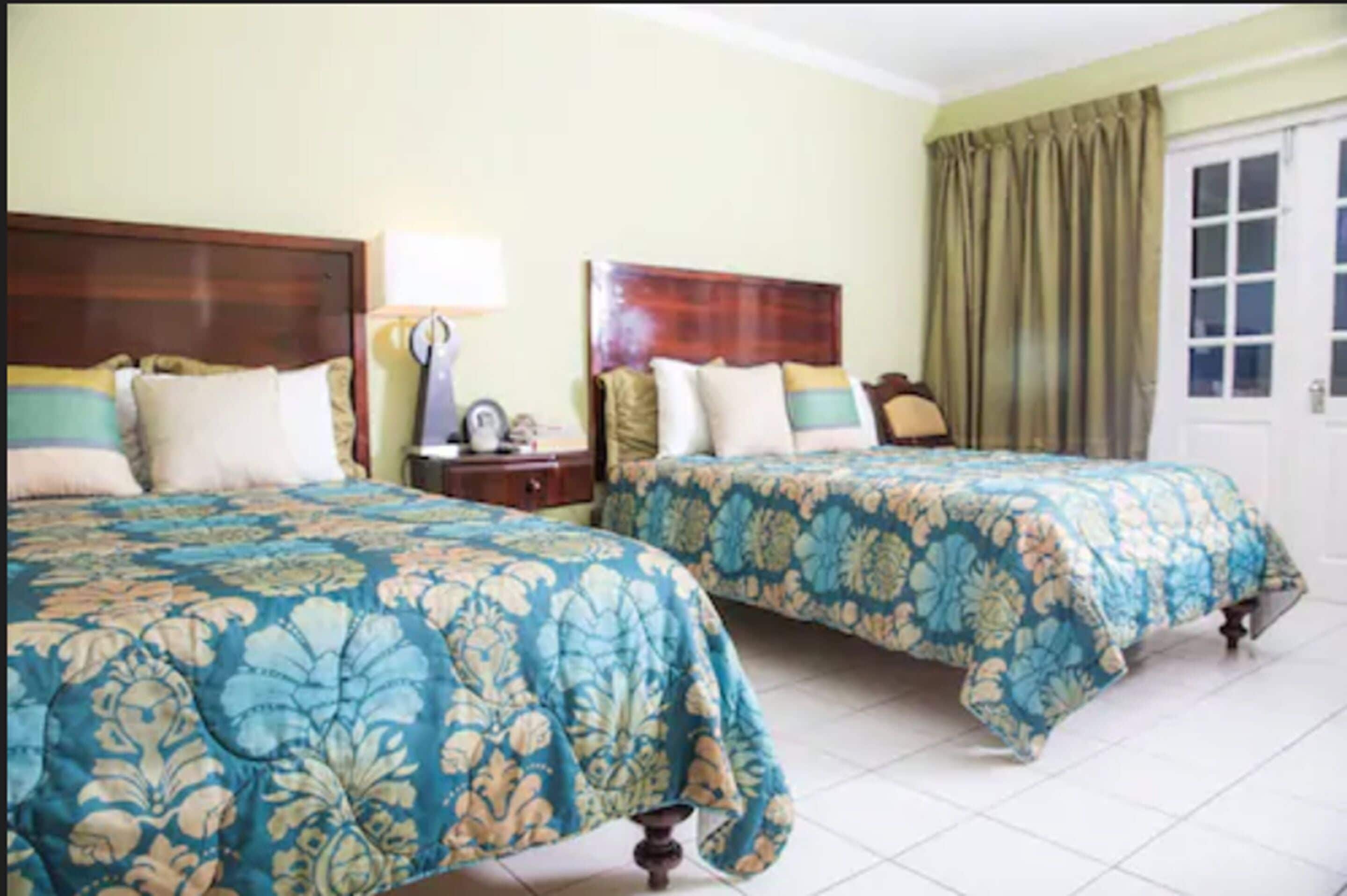 Une chambre d’hôtel avec deux lits, chacun recouvert d’une couvre-lit à motifs floraux, dotée d’une tête de lit en bois, d’une table de chevet et d’une porte avec des rideaux.