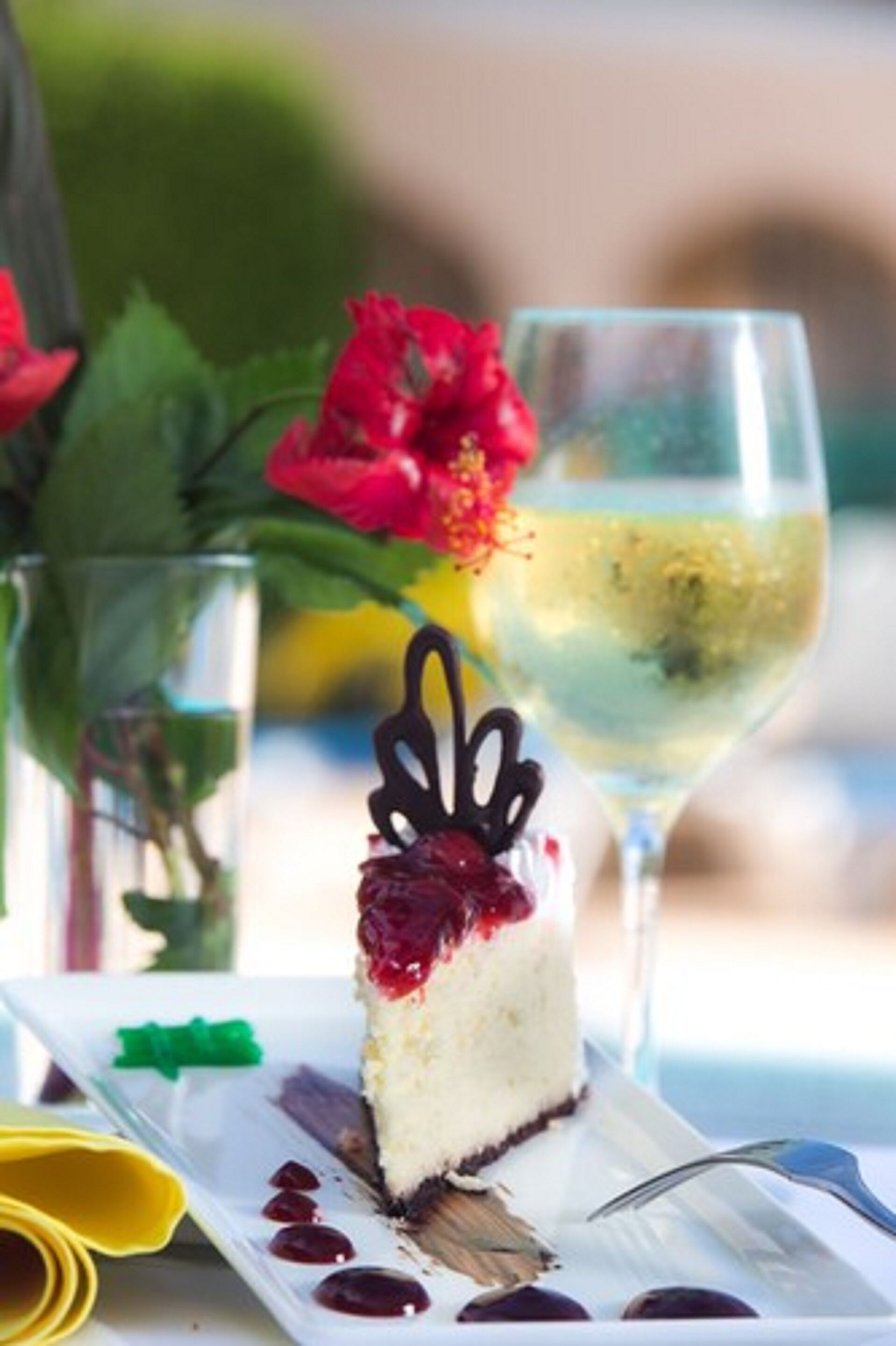 Uma fatia de cheesecake com cereja por cima, um copo de vinho branco e uma flor vermelha.