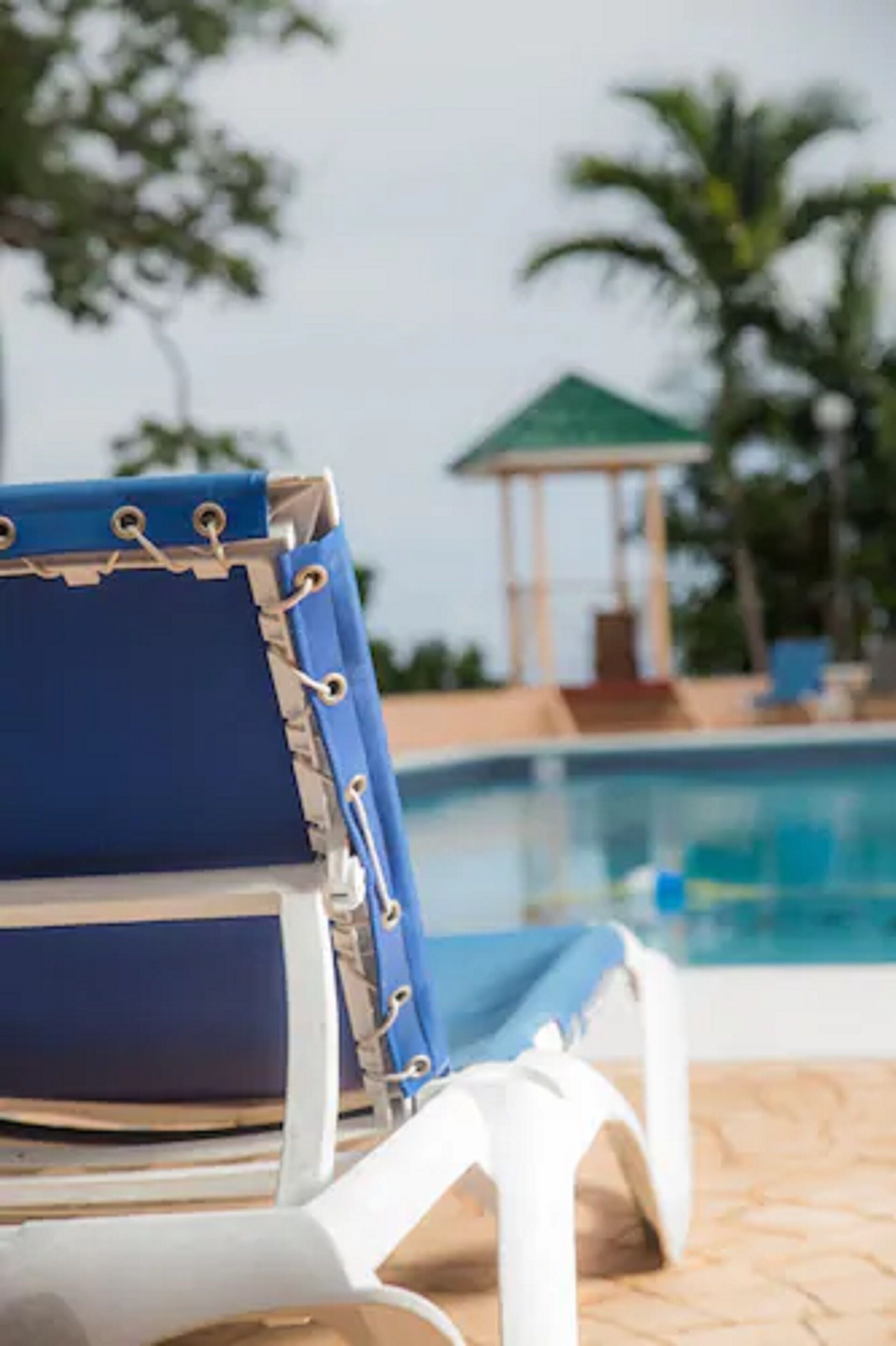 Ein blauer Liegestuhl mit weißen Armlehnen und einem weißen Kissen, gelegen in der Nähe eines Pools mit einer Laube im Hintergrund.