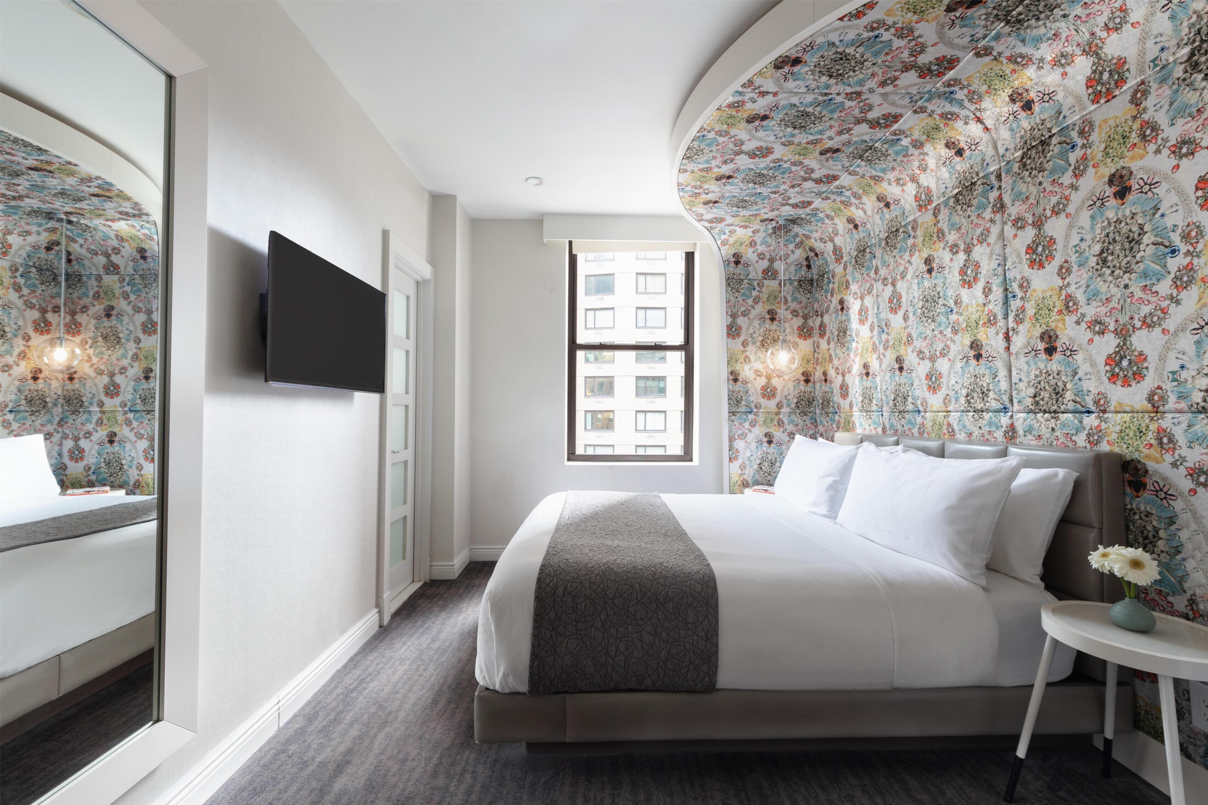 Una habitación de hotel moderna con una cama grande, un televisor de pantalla plana, una ventana con vista a edificios y papel tapiz floral.
