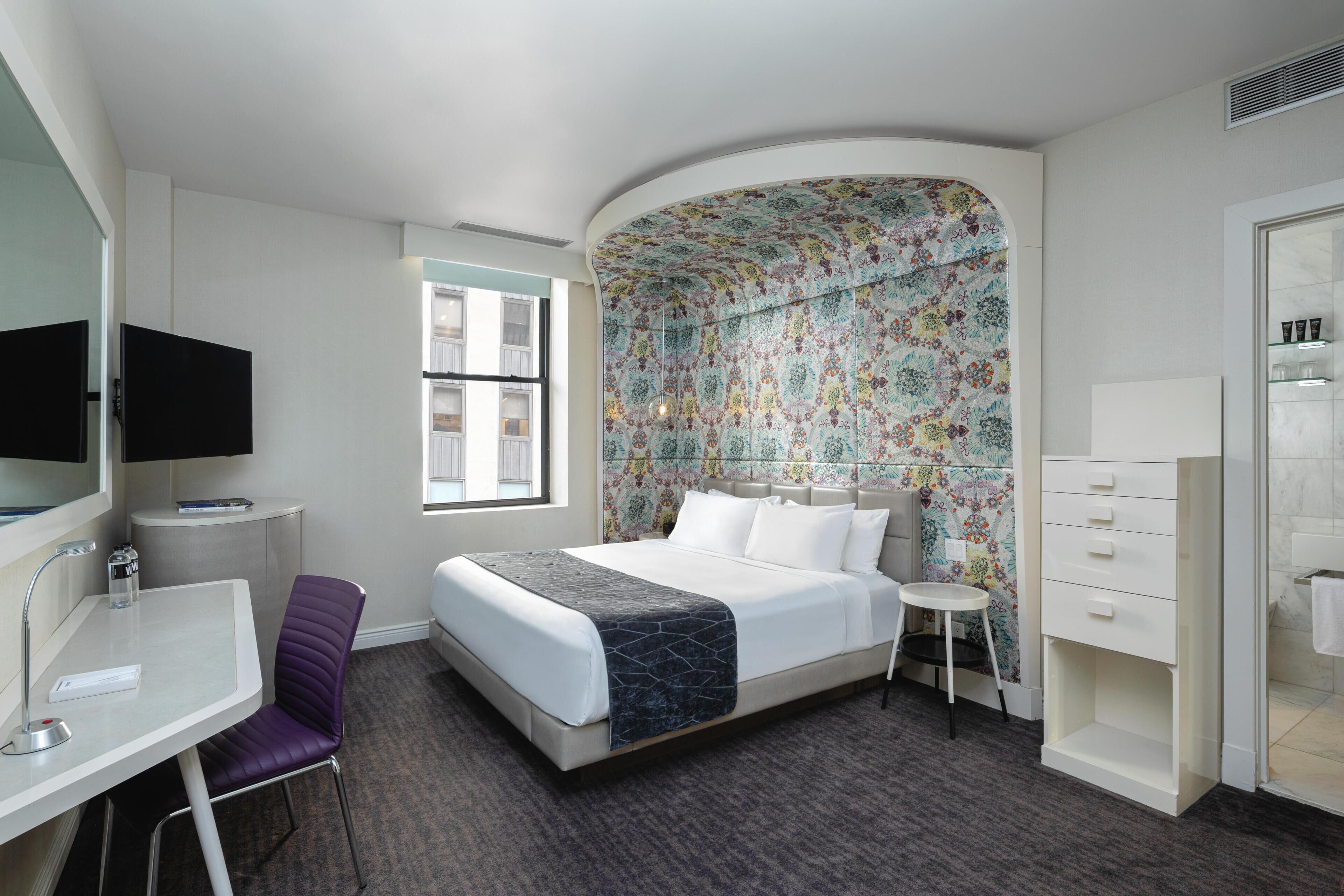 A hotel room with a bed, a desk, a chair, a small table, a mirror, and a shower area.