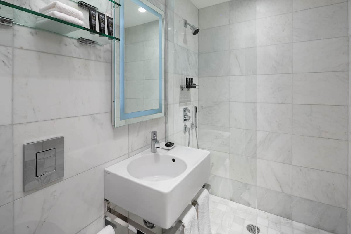 Un bagno moderno con un lavandino bianco, uno scaffale in vetro con articoli da toilette, una doccia e uno specchio.