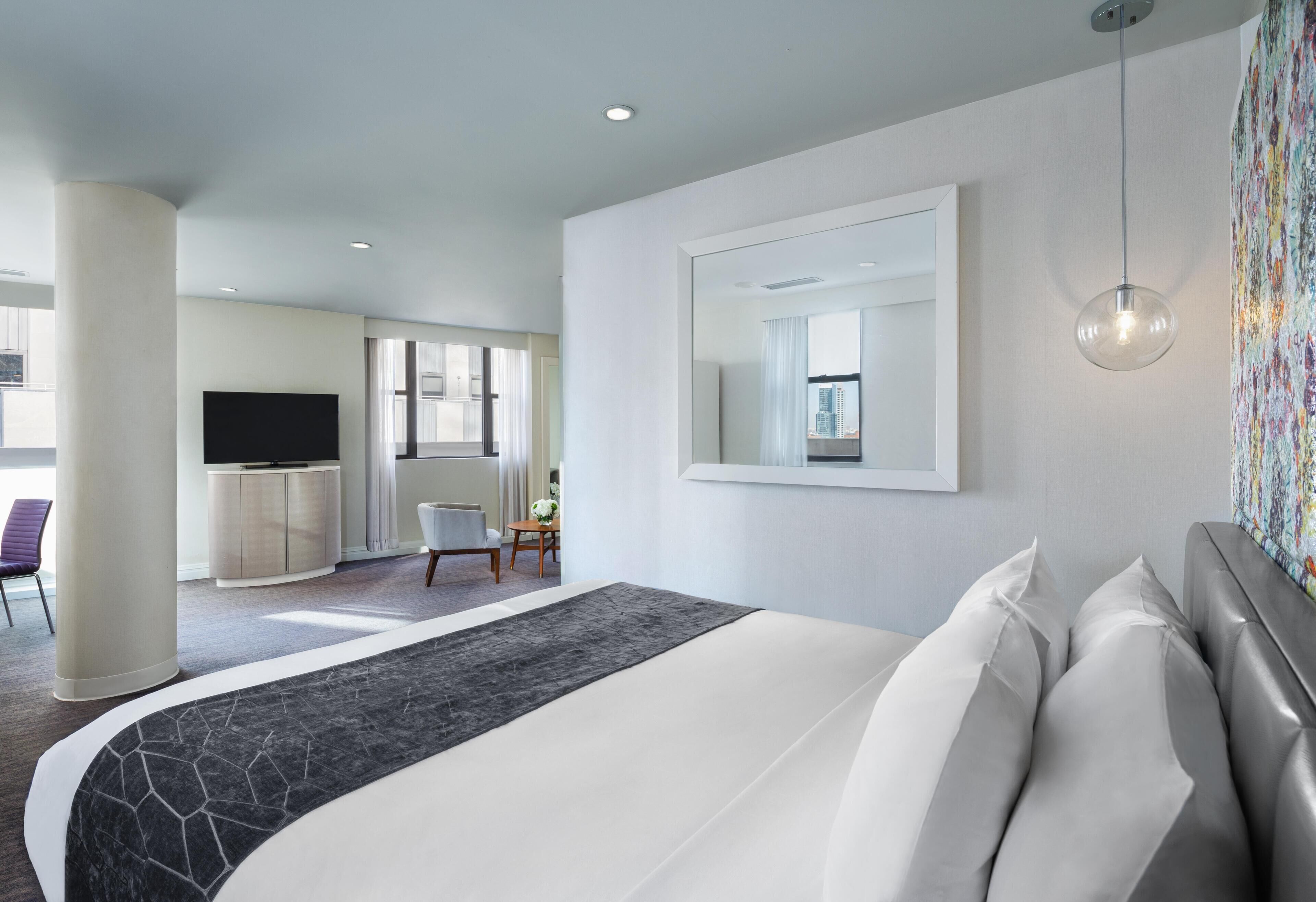A modern hotel room with a large bed, a flat-screen TV, a desk, and a dining area.