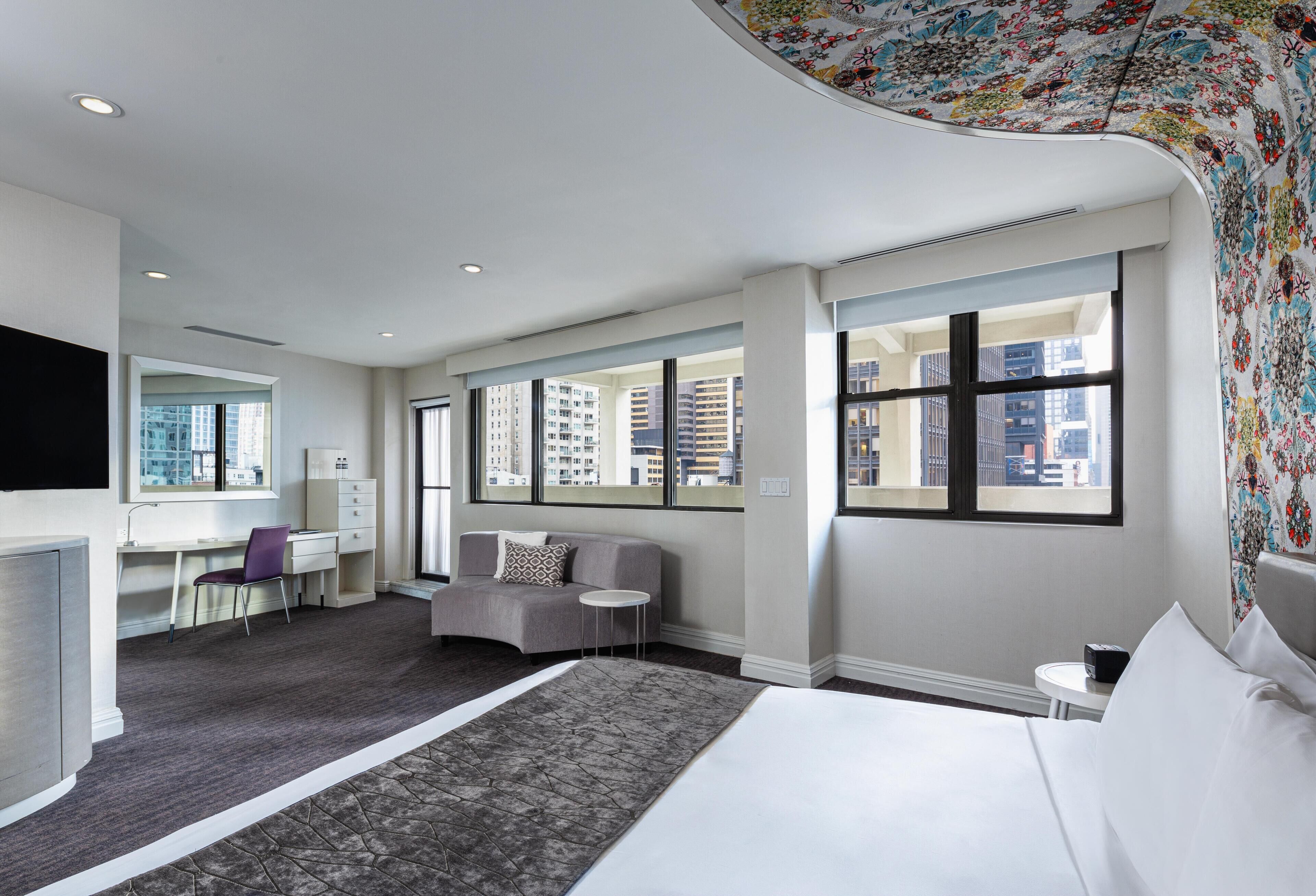 Een hotelkamer met een bed, een bureau, een stoel, een bank en grote ramen met uitzicht op de stad.