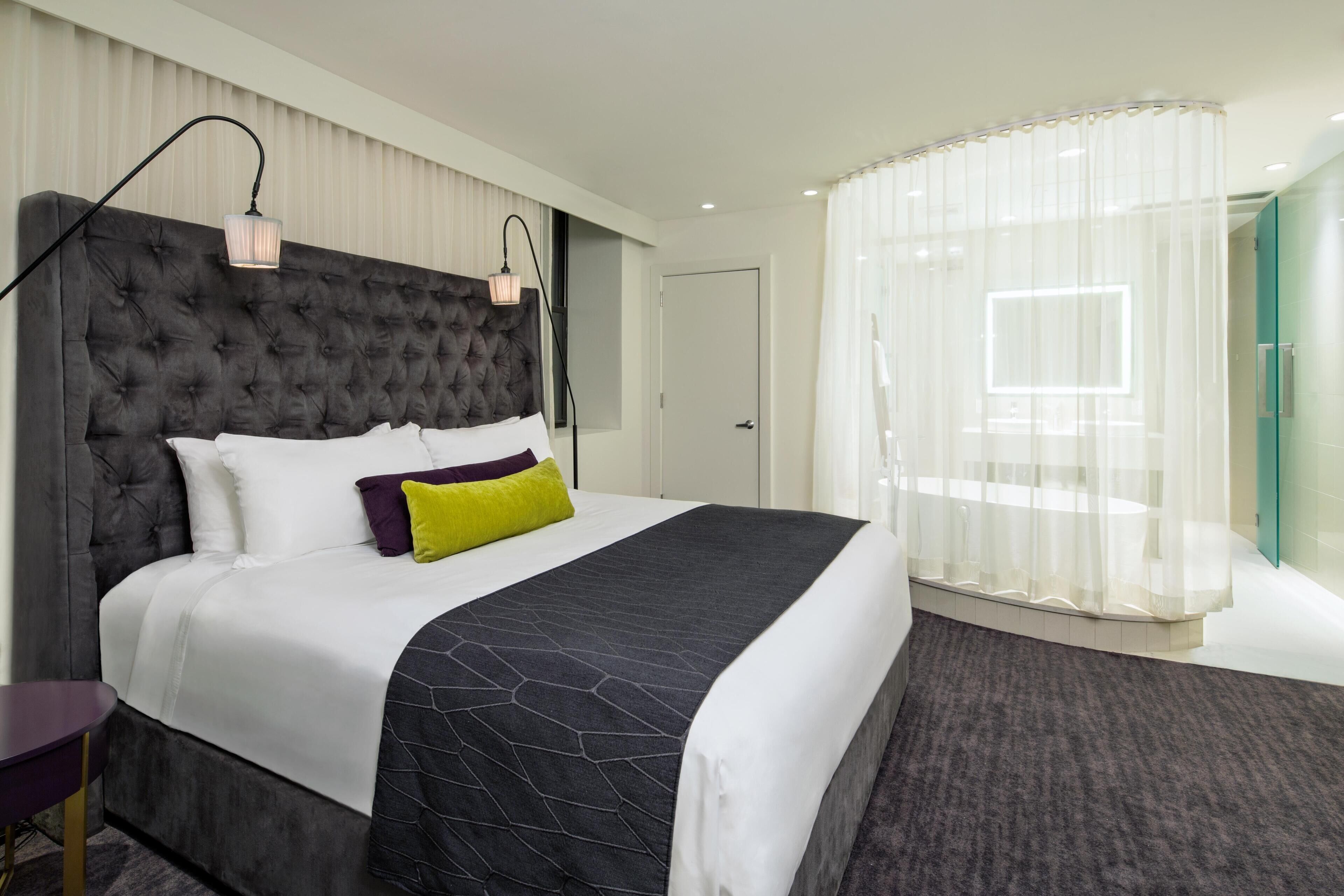 Una habitación de hotel moderna con cabecera capitonada, una cama grande con ropa de cama blanca y oscura, una mesita de noche y un panel de vidrio con vistas.