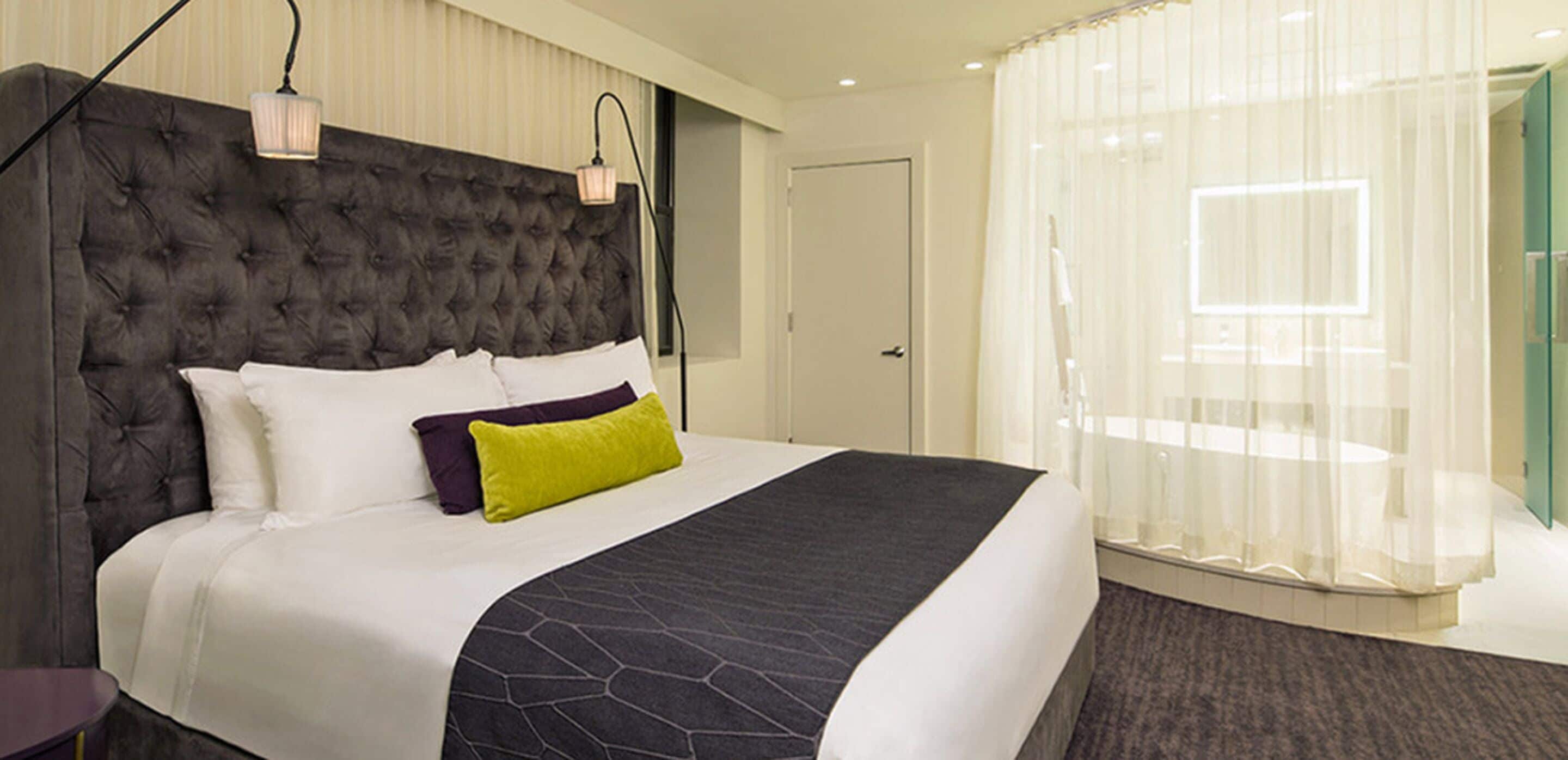 A large bed with a tufted headboard, white bedding, and a dark patterned throw. There is a small bedside table and a lamp on the wall. The room has a large window with sheer curtains.