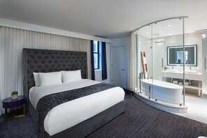 A modern hotel room with a large bed, a glass-enclosed shower, a separate bathroom with a large mirror, and a small side table with a plant. - Dream Midtown, by Hyatt (New York)