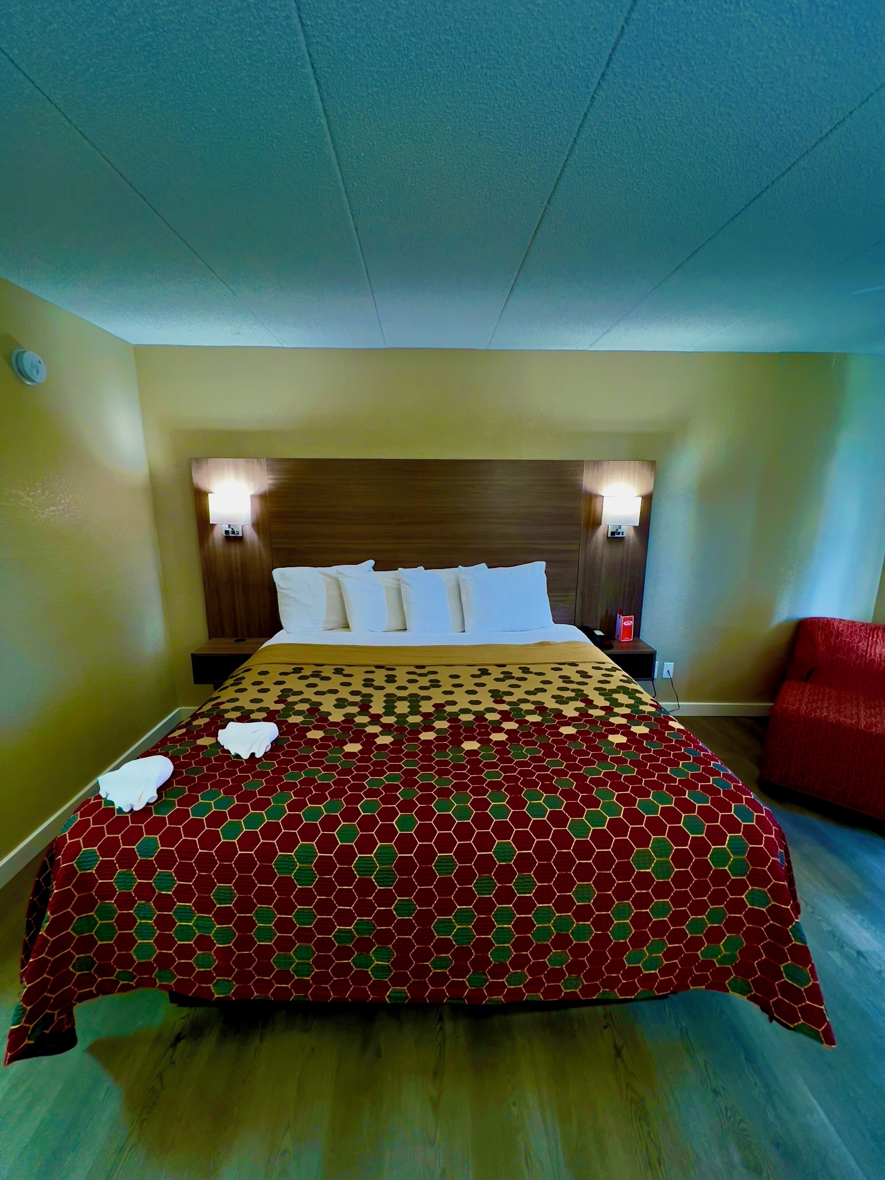 Quarto de hotel com cabeceira de madeira, duas mesas de cabeceira com luminárias, colcha de cama estampada em vermelho e uma poltrona vermelha.