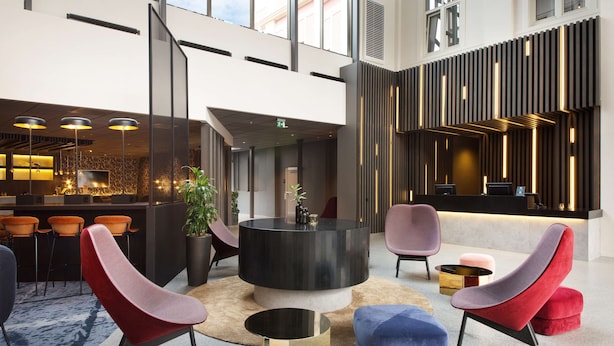 Een moderne lobby met een ronde, zwarte salontafel, rode en blauwe stoelen en een receptiebalie.