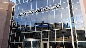 Een modern gebouw met grote glasramen en de tekst 'SCANDICAMBASSADEUR DRAMMEN' op de gevel.