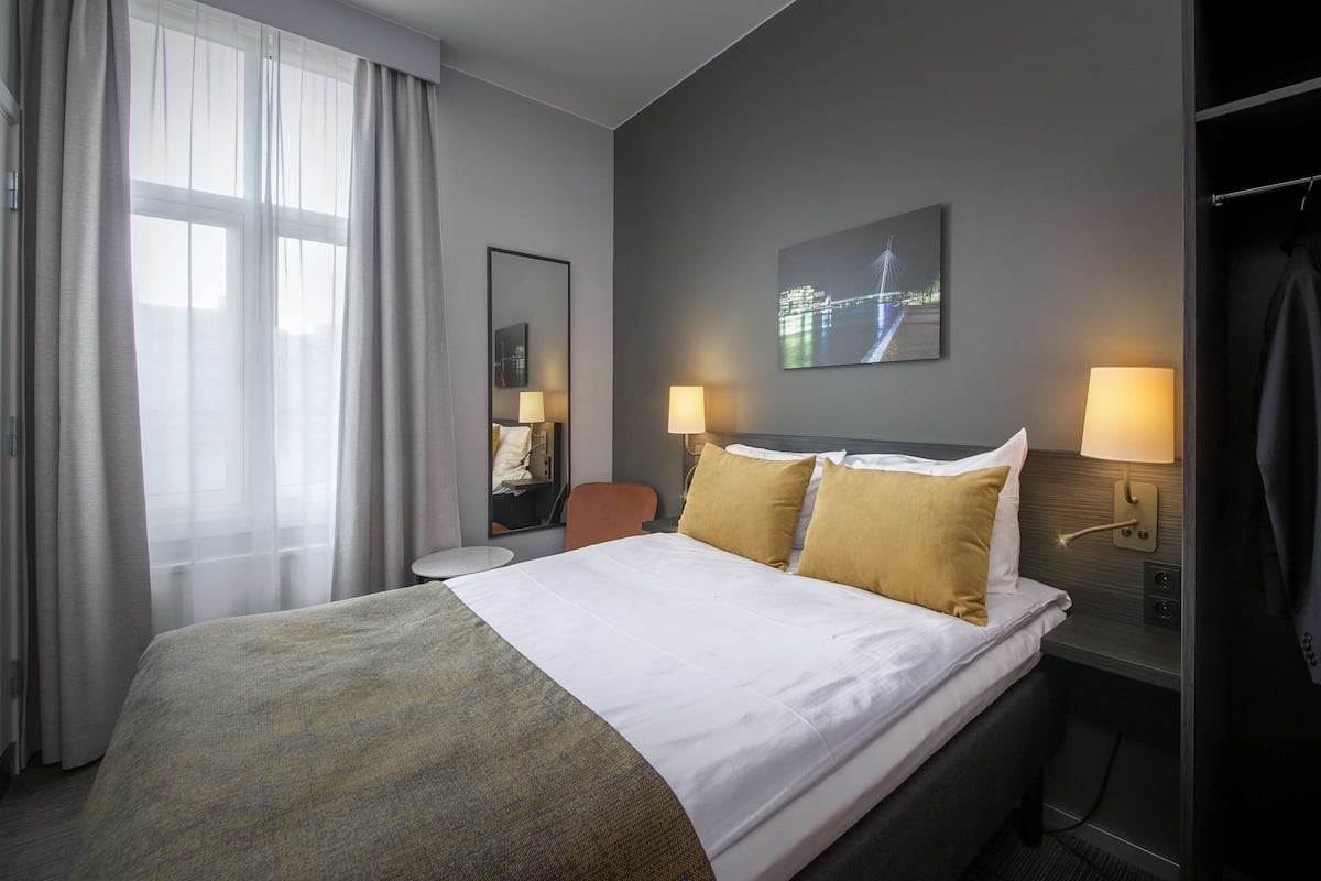 Una habitación de hotel con una cama grande, dos lámparas de noche, una mesita de noche, una silla, un espejo y una ventana con cortinas.