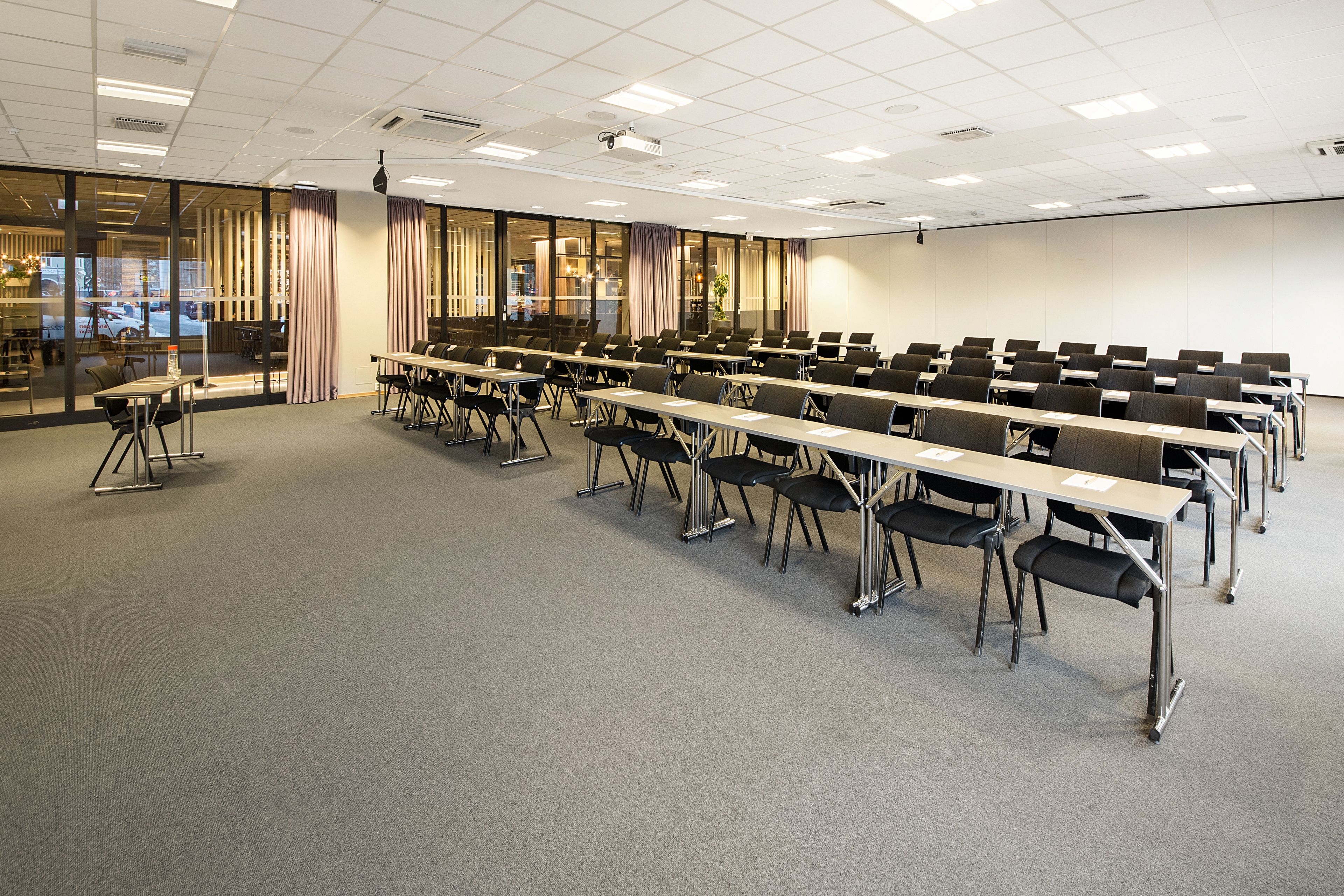 Un salón de conferencias con filas de mesas y sillas, ventanas amplias con cortinas y un proyector en el techo.