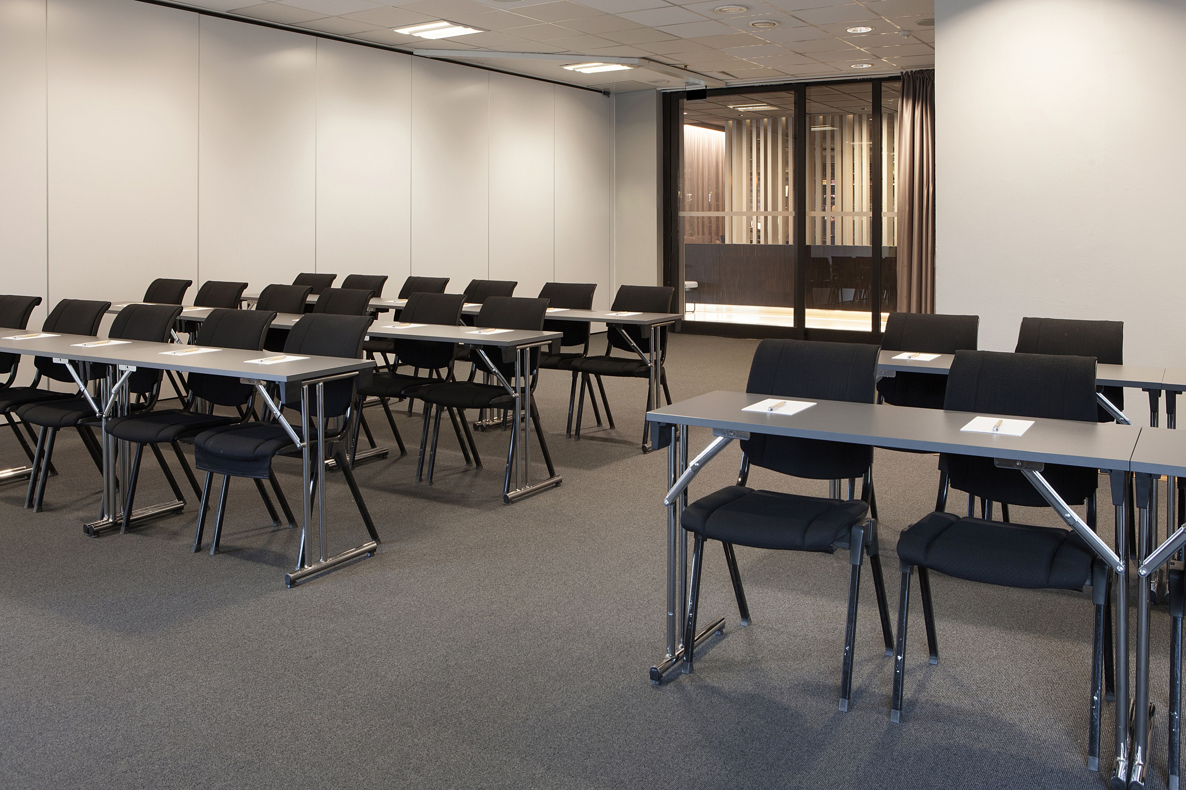 A conference room with rows of tables and chairs, a large screen, and a glass partition with curtains.