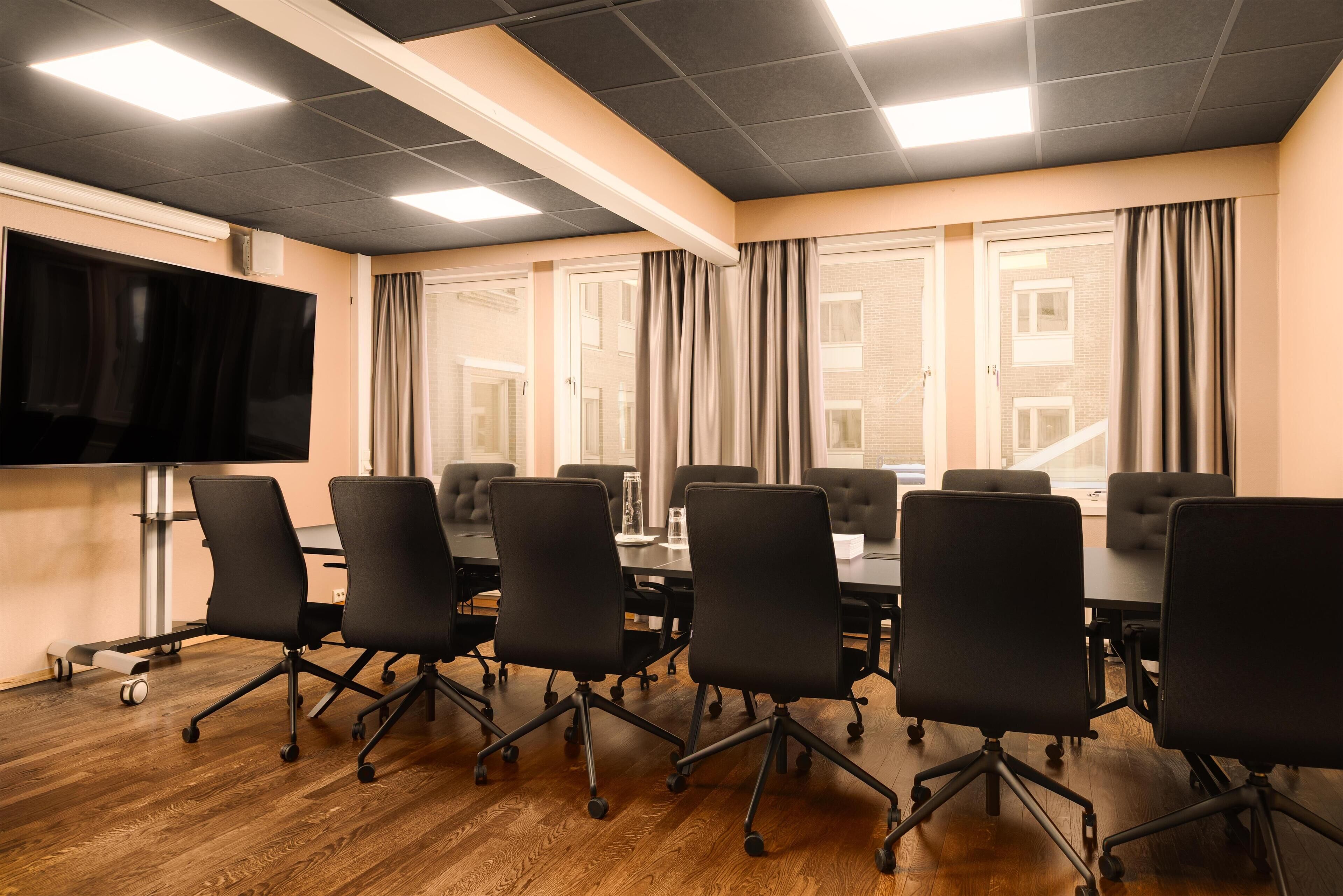 Un salón de conferencias con una mesa grande, sillas de oficina negras, un televisor de pantalla plana y ventanas amplias con cortinas.