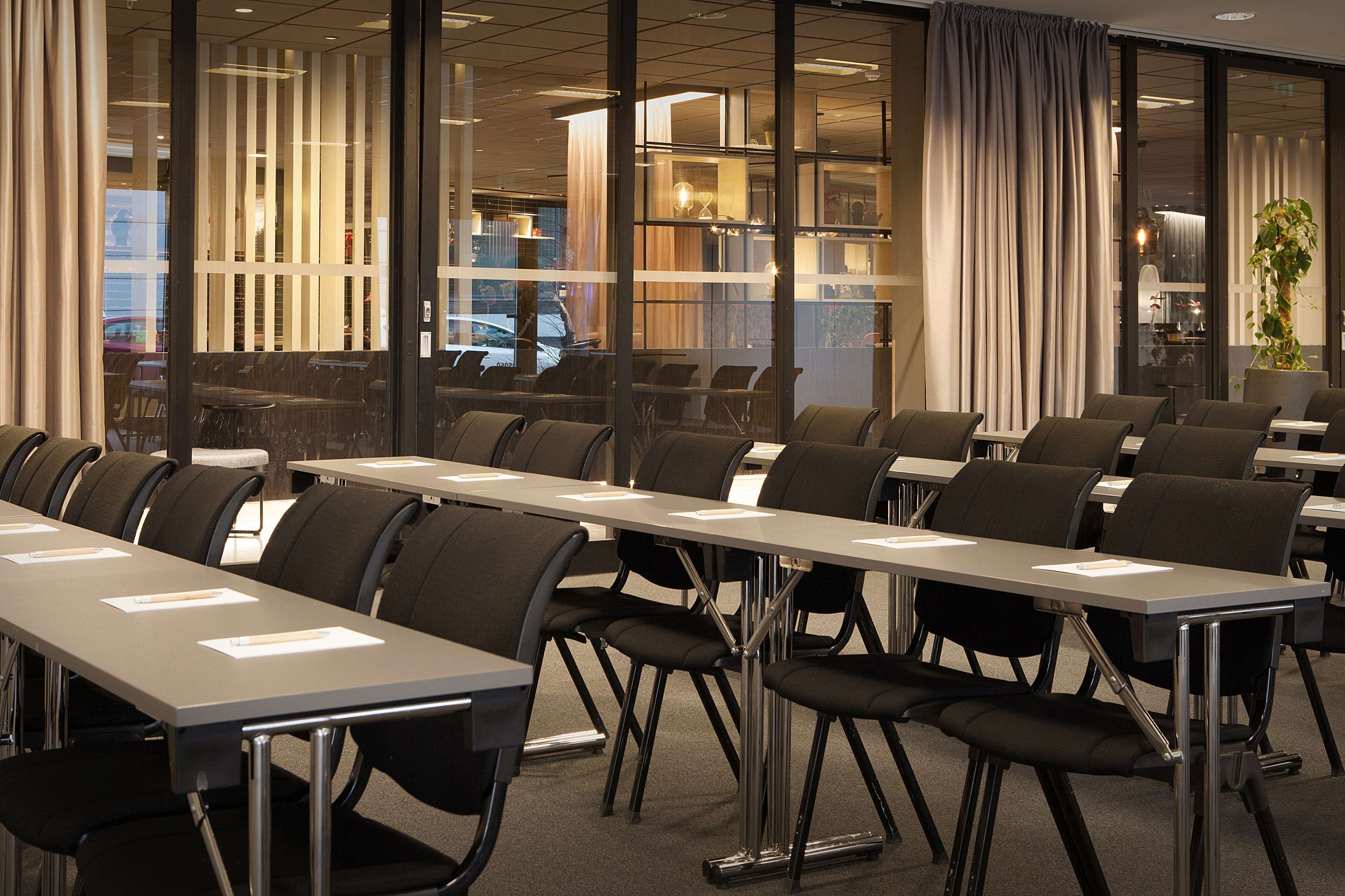 Un salón de conferencias con varias mesas y sillas, ventanas amplias con cortinas y una planta en la esquina.
