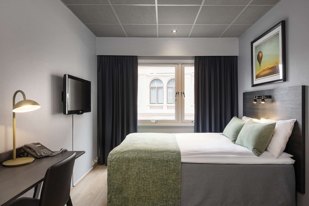 Una habitación de hotel con cama, escritorio, teléfono, televisión y ventana con cortinas.
