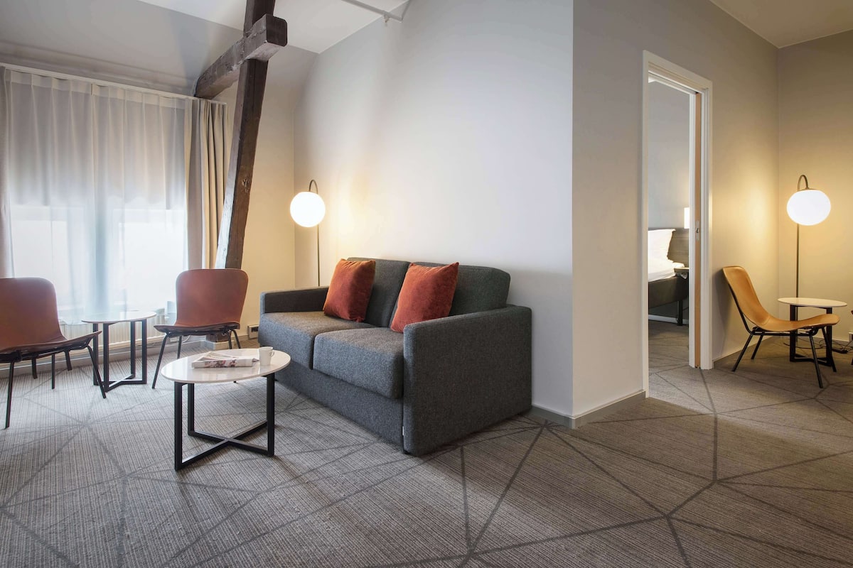 Una habitación de hotel moderna con un sofá gris, cojines rojos, una mesita blanca pequeña y dos sillas. Hay dos lámparas de pie y un ventanal con cortinas traslúcidas.
