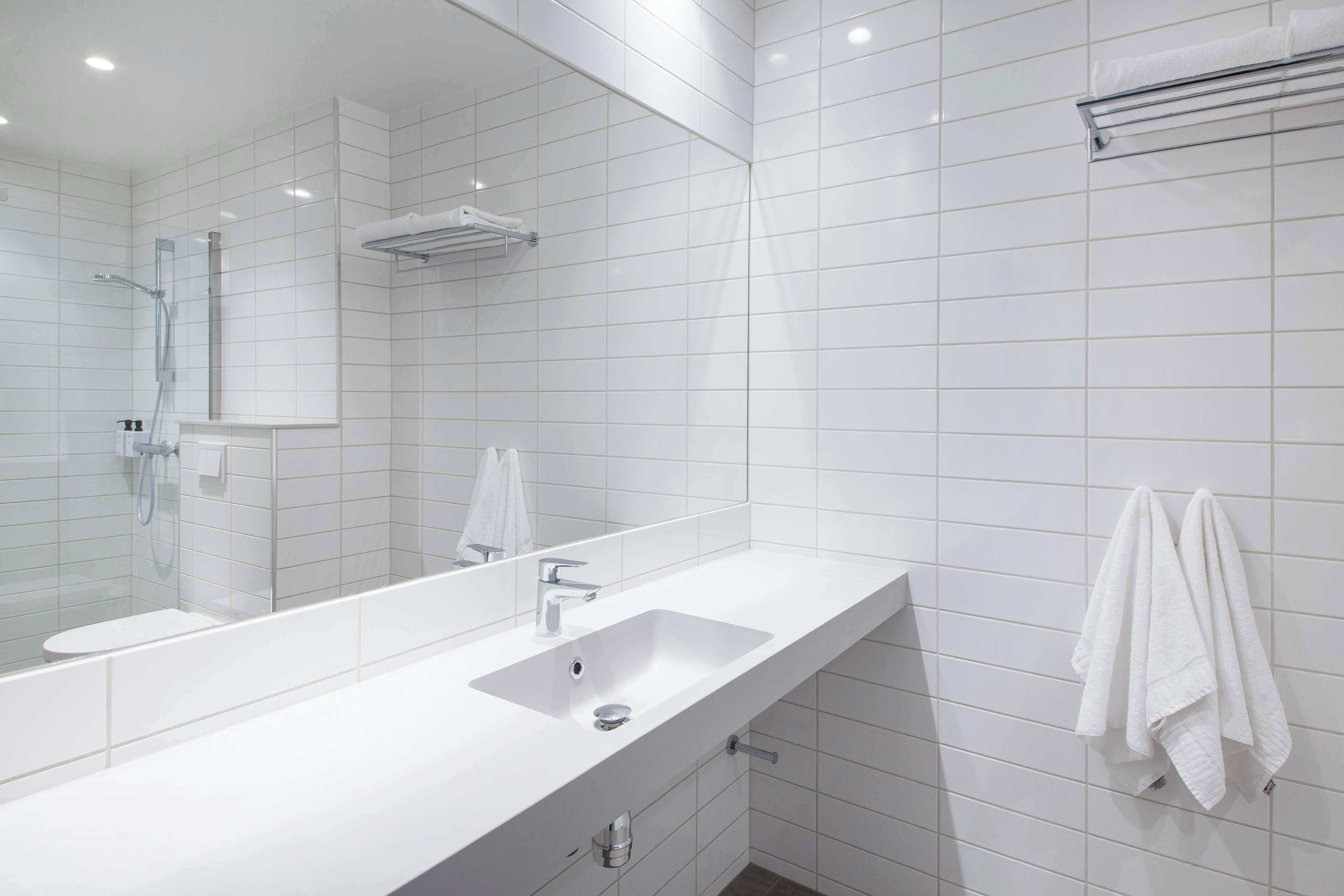 A modern bathroom with white tiles, a large mirror, a sink, and a towel rack.