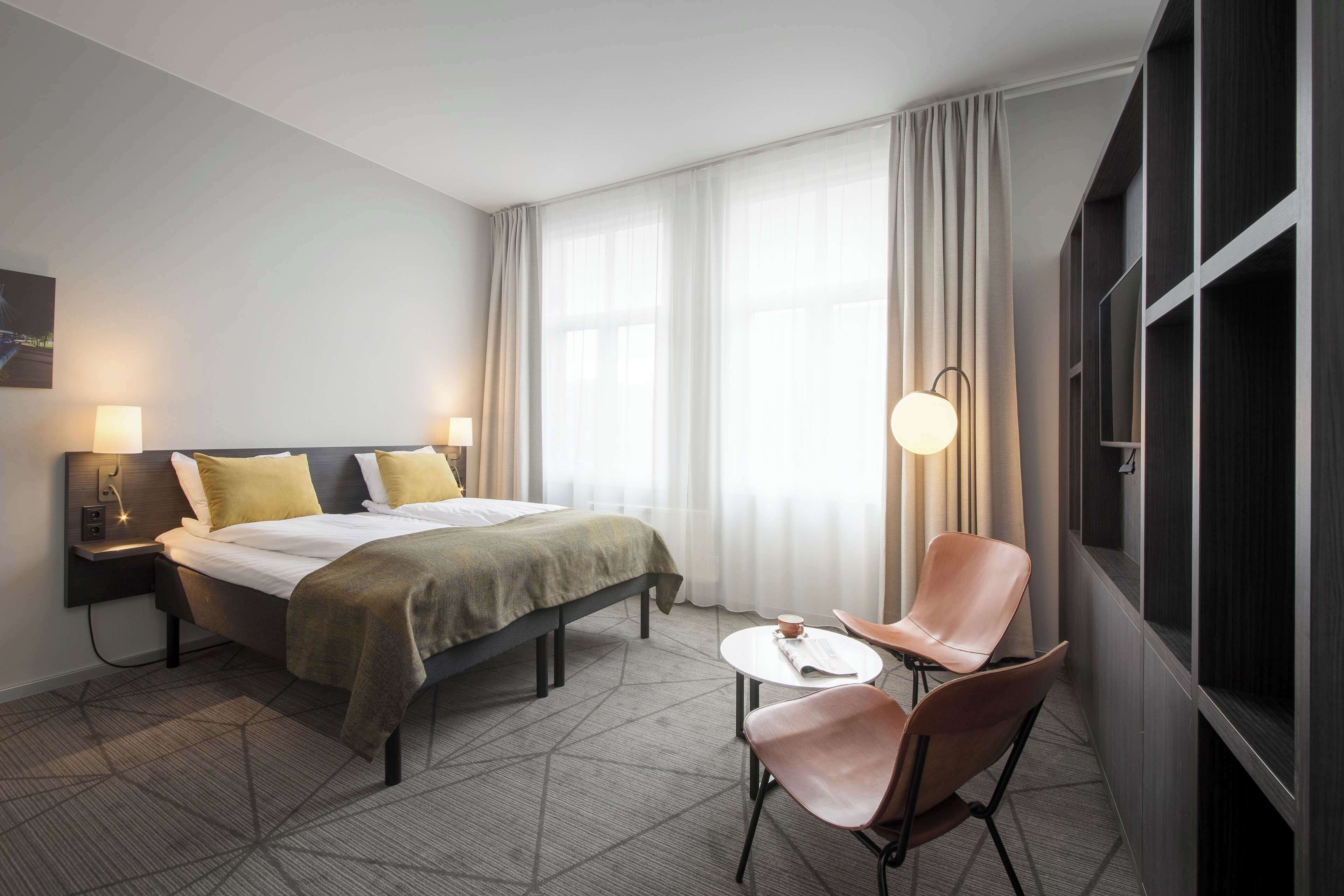 A hotel room with a bed, bedside tables, a chair, a small table, and a window with curtains.