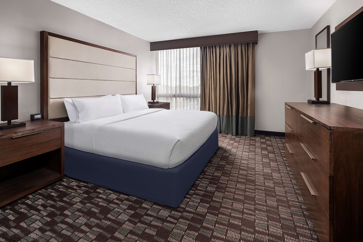 Ein Hotelzimmer mit einem großen Bett, einem Holznachttisch, einem Schminktisch, einem Flachbildfernseher und einem Fenster mit Vorhängen.