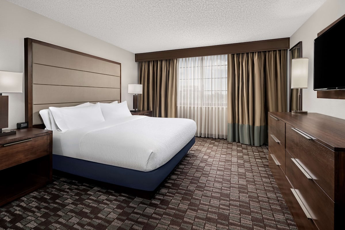 Ein Hotelzimmer mit einem großen Bett, einem Holzkopfende, einem Schminktisch, einem Flachbildfernseher und einem Fenster mit Vorhängen.
