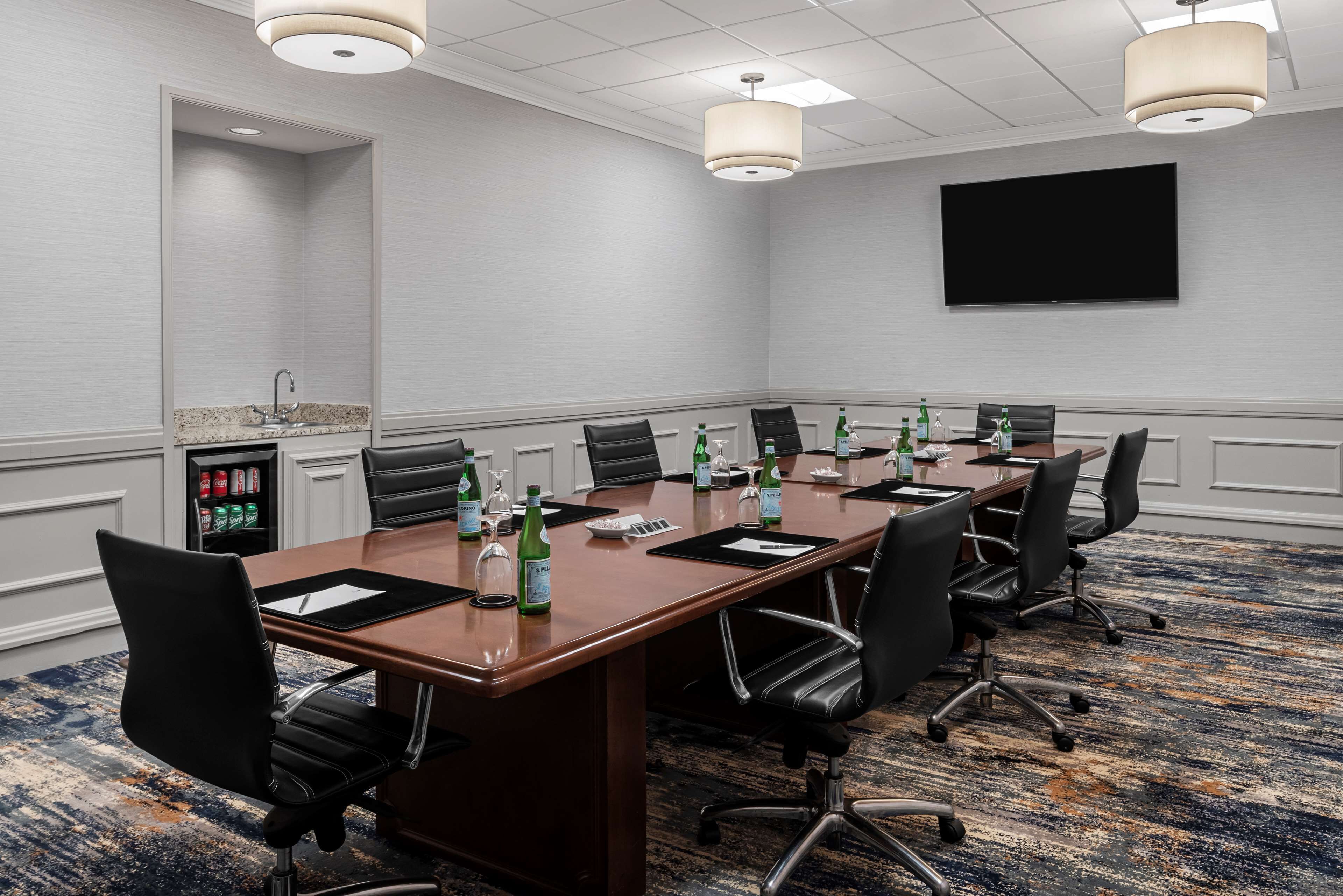 Una sala de conferencias con una mesa larga, varias sillas, un televisor de pantalla plana y un minibar con bebidas.