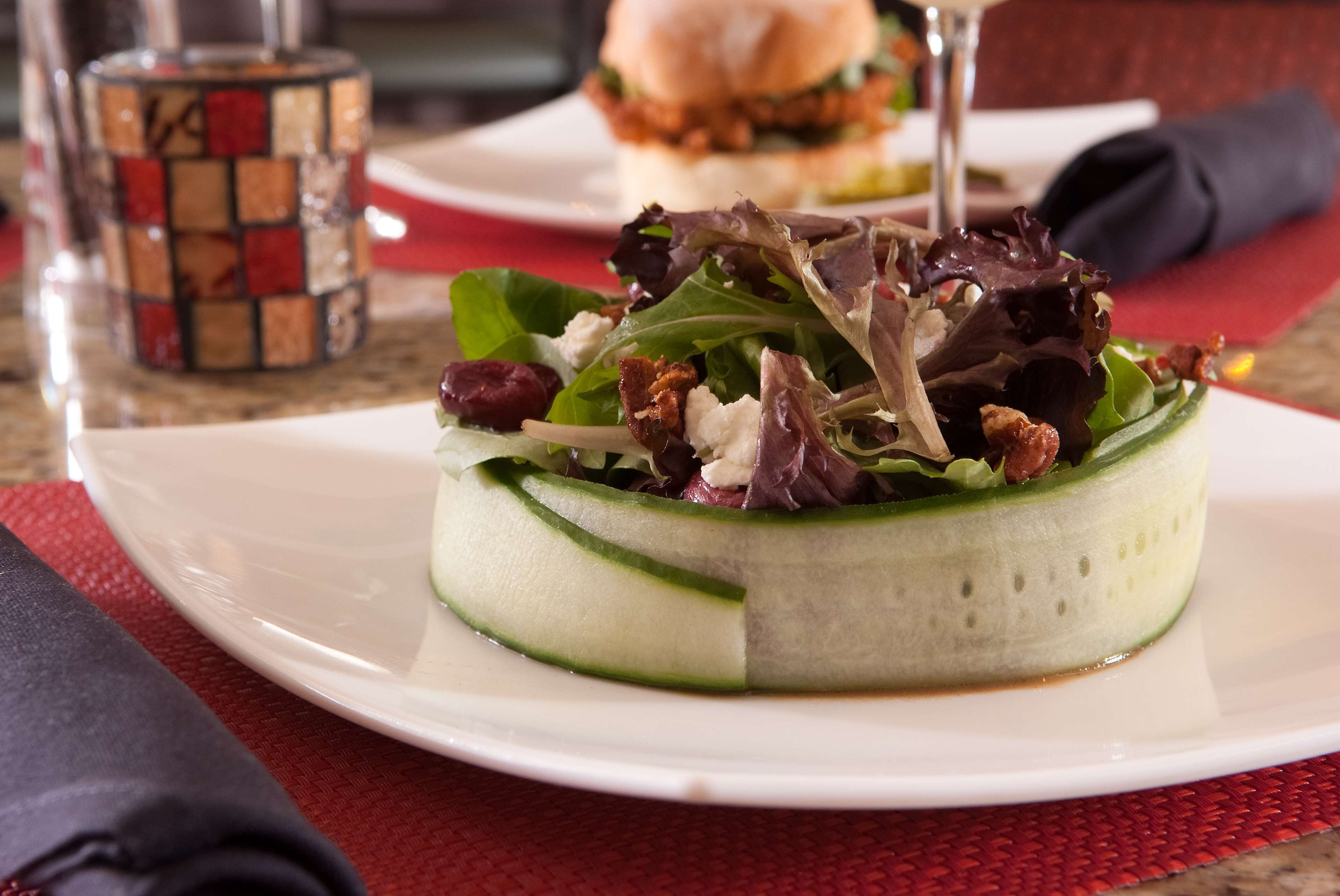 A salad on a white plate with a red placemat, a glass with a mosaic pattern, and a sandwich in the background.