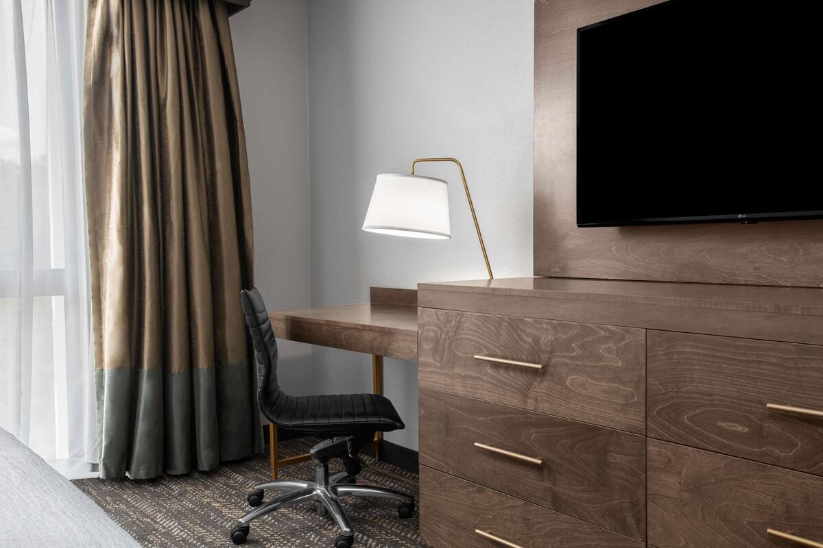 Ein Hotelzimmer mit einem Holztisch, einem schwarzen Bürostuhl, einem flachbildfernseher an der Wand und einer Lampe auf dem Tisch.