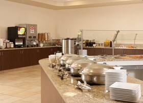 Uma mesa de buffet com diversos recipientes de comida, uma máquina de café e pilhas de pratos.