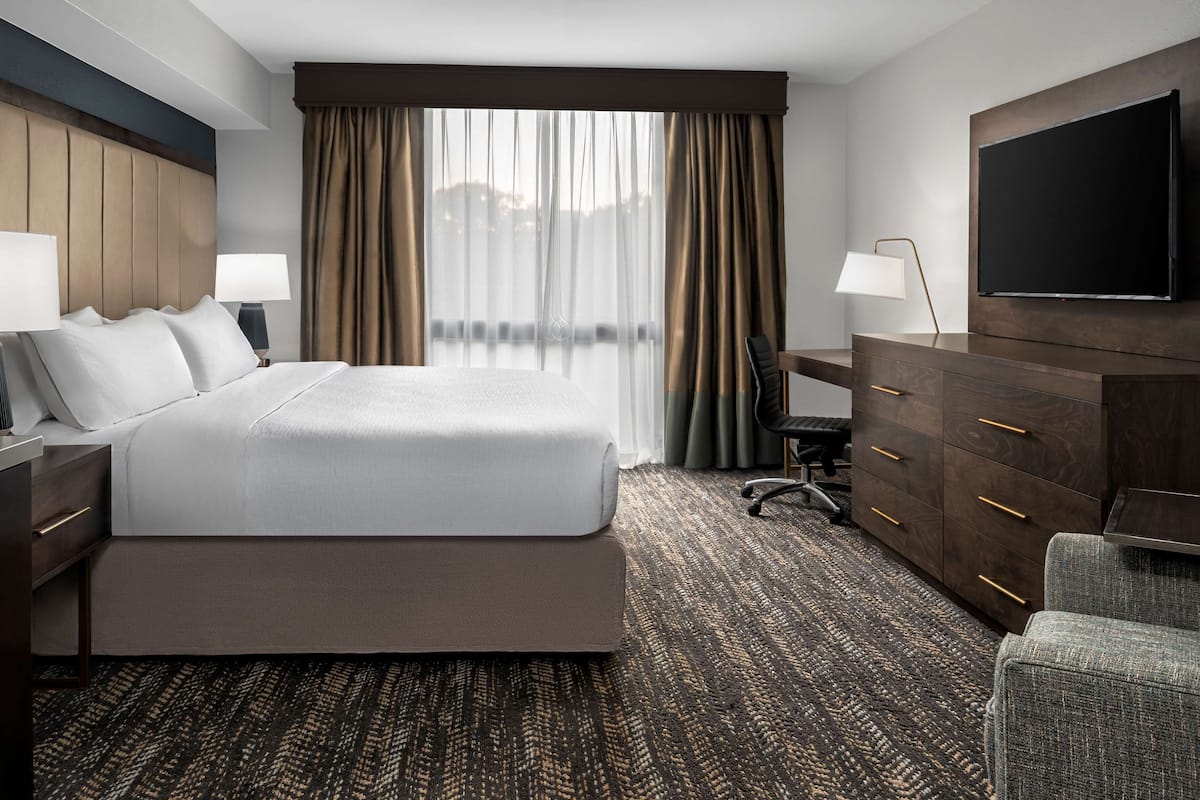 Ein Hotelzimmer mit einem großen Bett, einem Schreibtisch mit Lampe, einem Schminktisch, einem Sessel und einem an der Wand befestigten Fernseher.