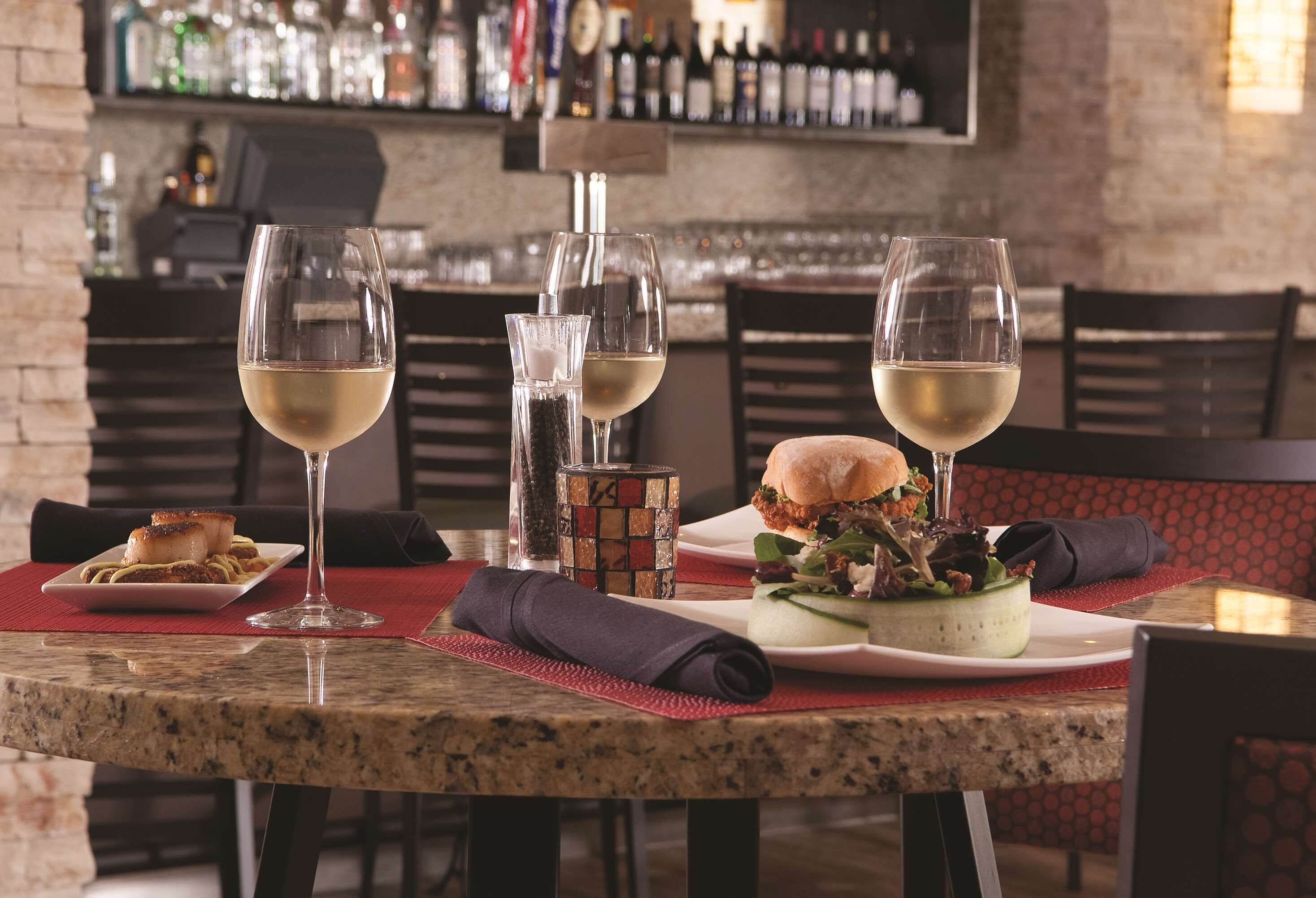 A restaurant setting with a bar in the background, a table with three wine glasses, a burger, and a salad.