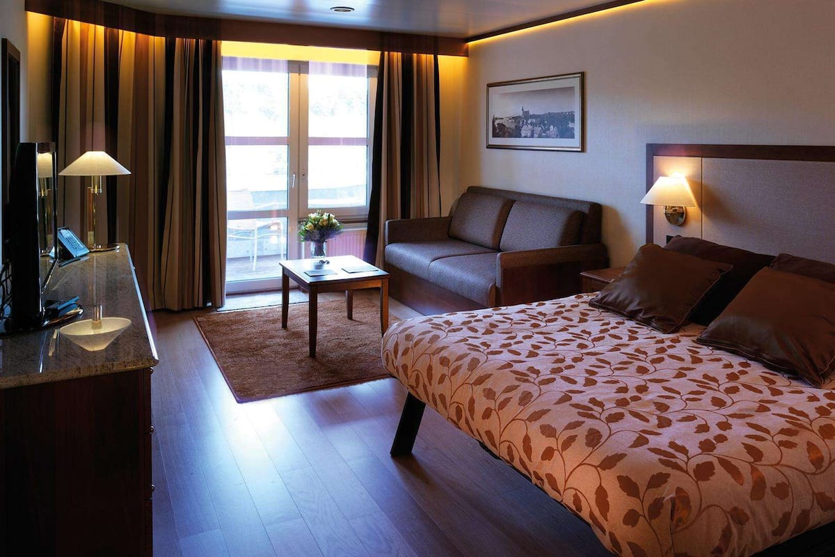A hotel room with a bed, a sofa, a desk with a lamp, and a view of the outdoors.
