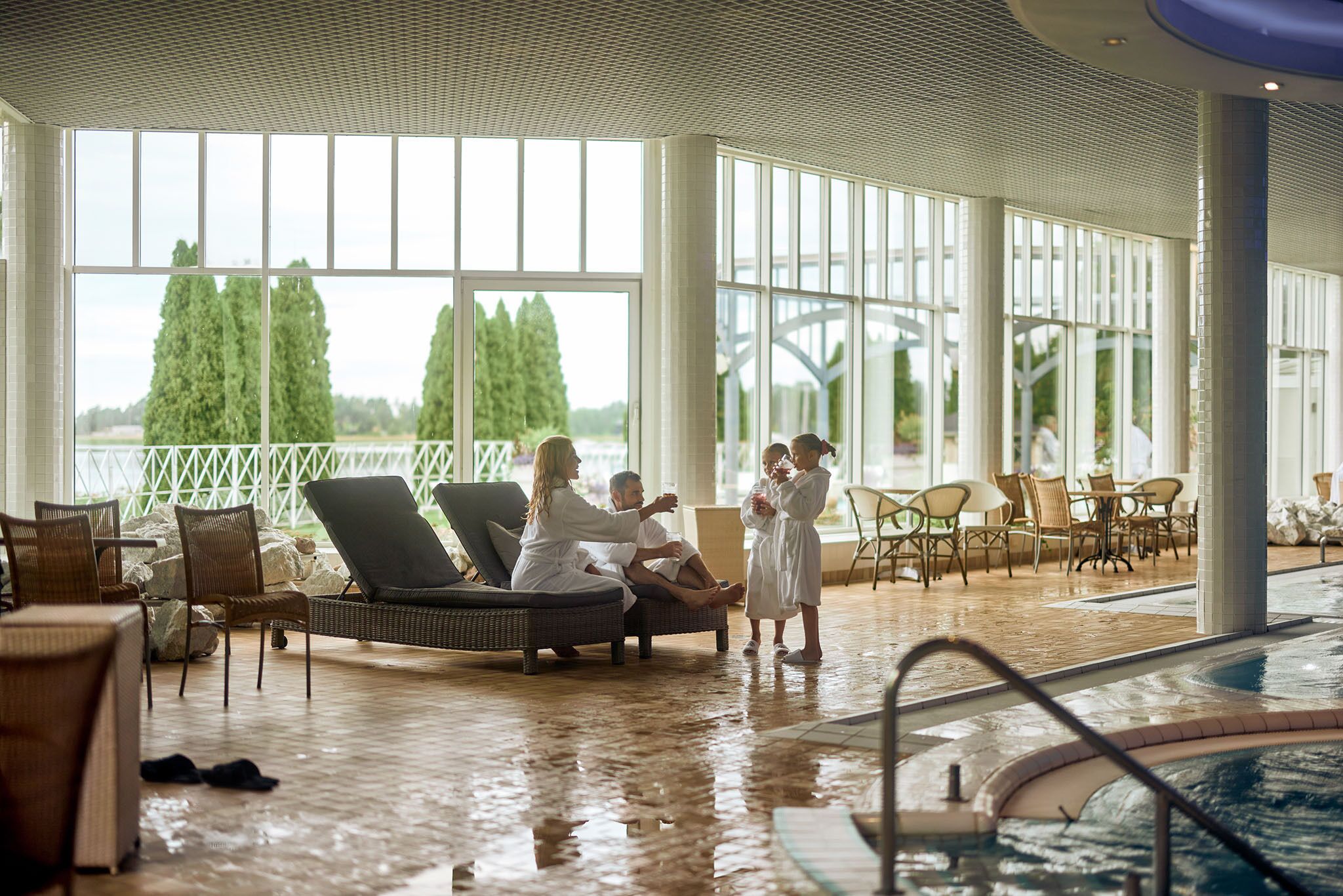 Uma área de piscina interior ampla, com espreguiçadeiras e mesa de bilhar, grandes janelas com vista para árvores e uma ponte, e um ambiente bem iluminado e limpo.