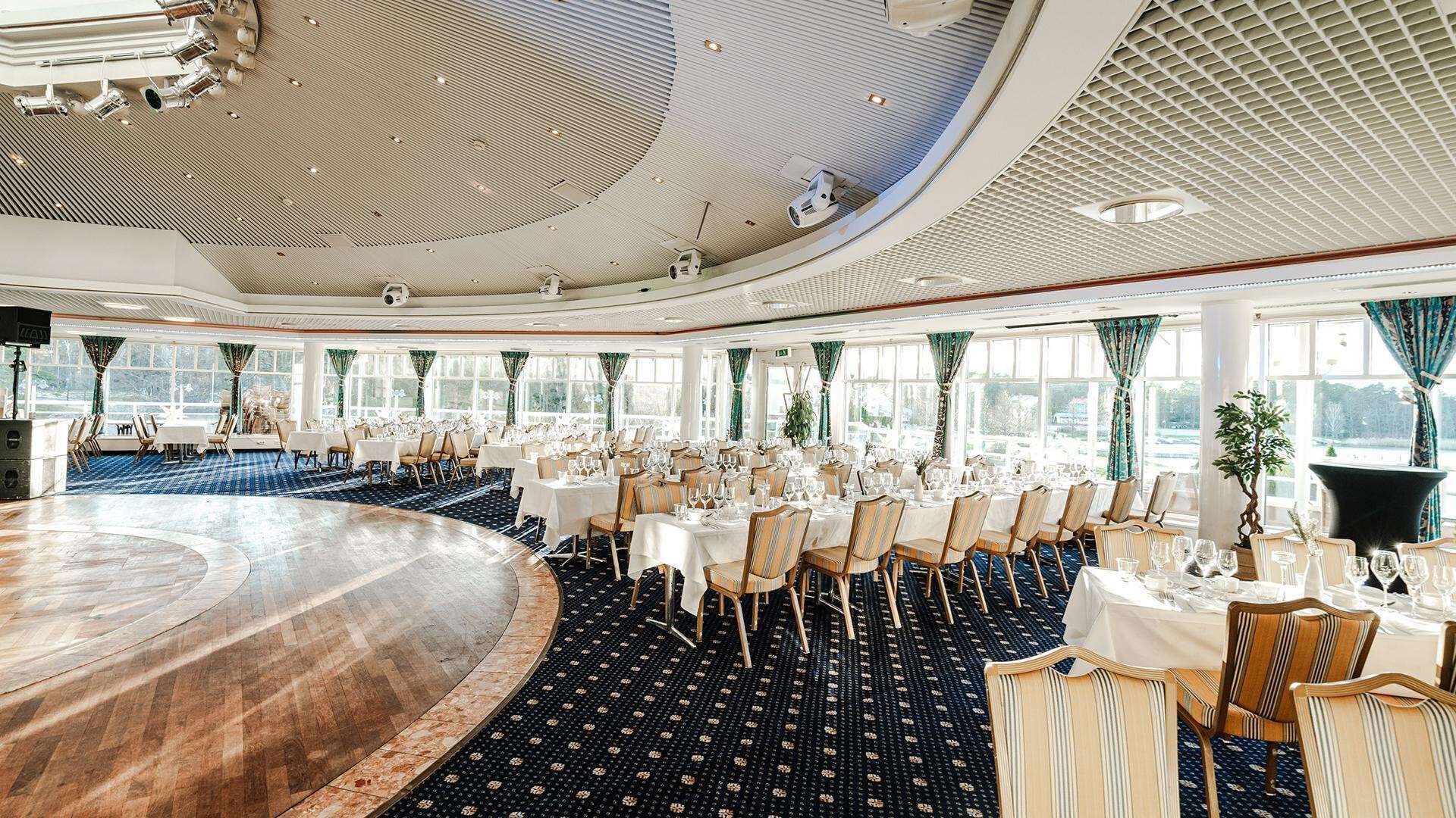 Un amplio y elegantemente decorado salón comedor con mesas redondas, sillas y una zona central elevada.