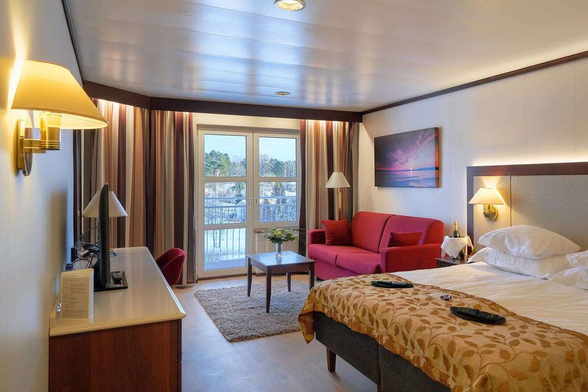 A hotel room with a bed, a red armchair, a desk with a lamp, and a view of the outdoors through a large window.